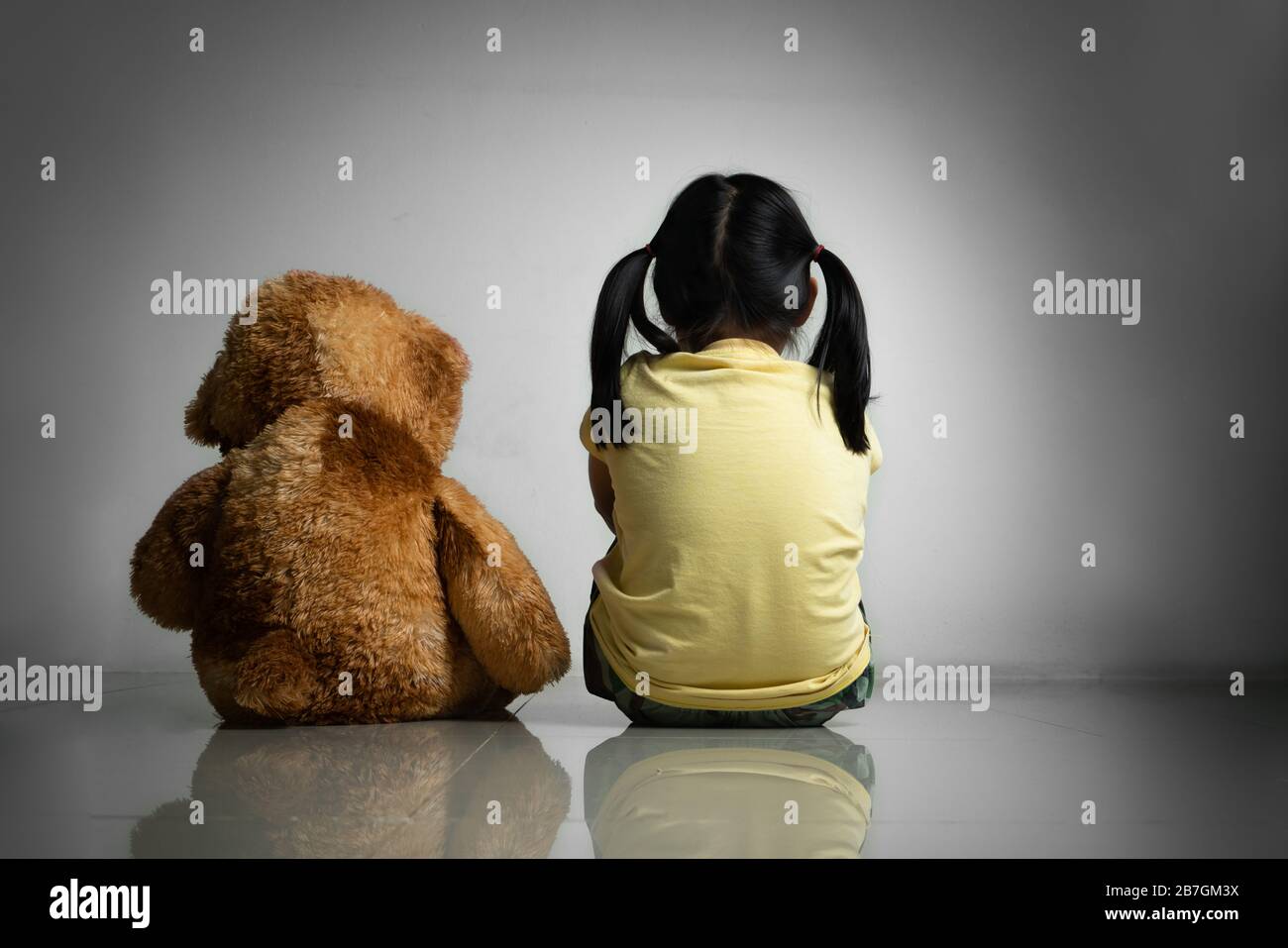 Little girl with teddy bear sitting on floor at empty room. Mental and depressed family concept. Back view Stock Photo