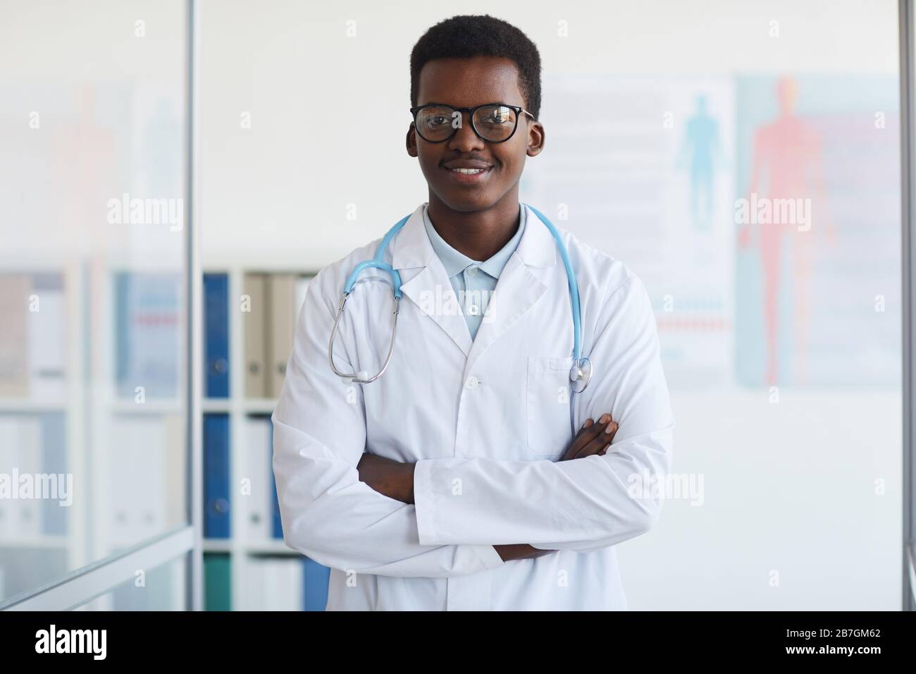Waist up portrait of young African-American doctor standing with arms crossed and smiling at camera while posing in med clinic, copy space Stock Photo