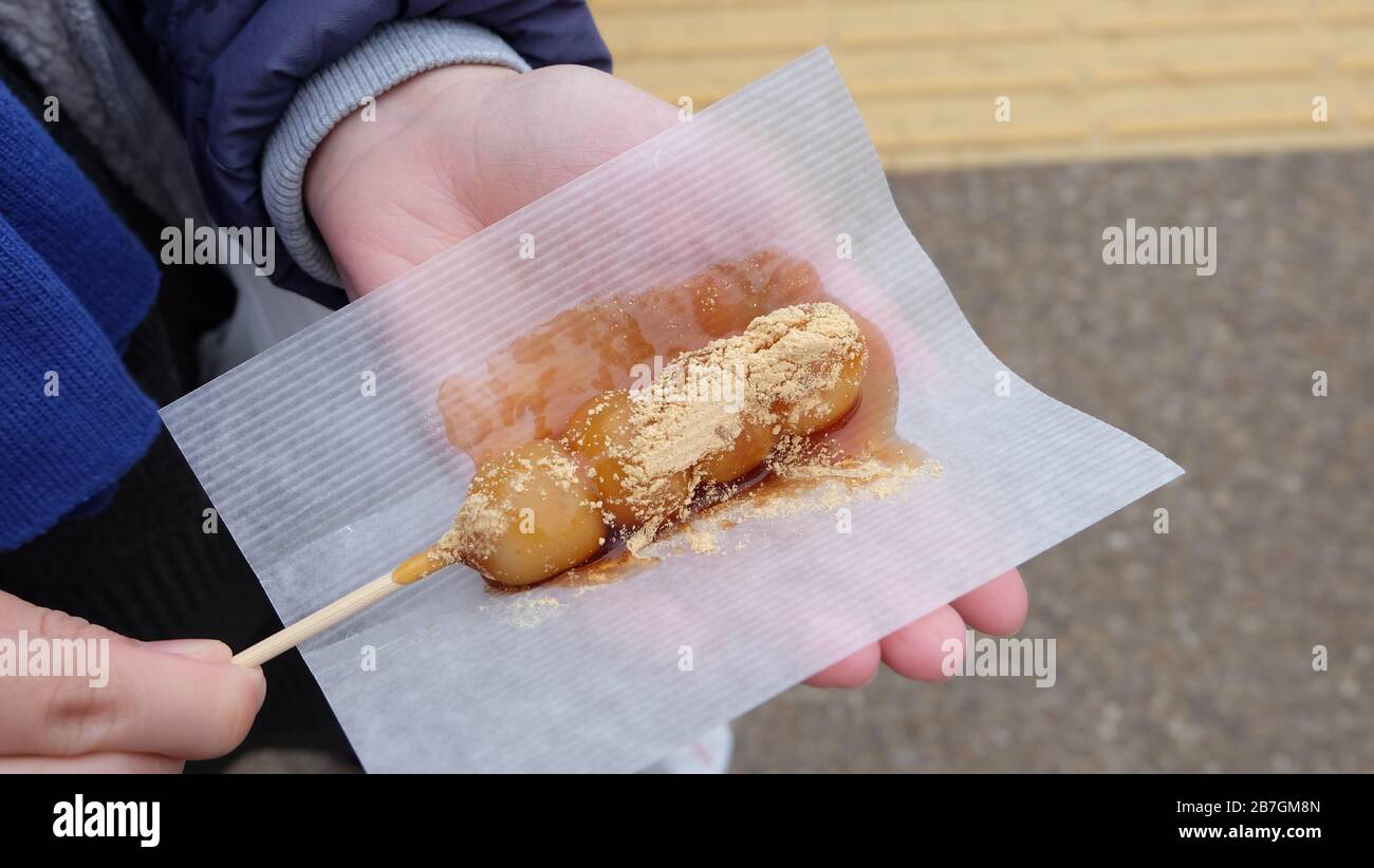 Hand holding a skewer of mitarashi dango sprinkled with kinako soy bean powder, on a piece of paper. Stock Photo