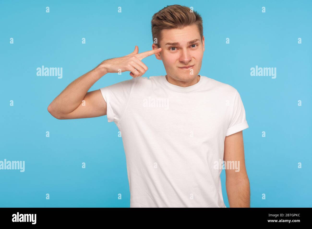 You are idiot. Displeased man in casual white t-shirt holding finger against his temple, making crazy stupid gesture, blaming in senseless dumb talk. Stock Photo