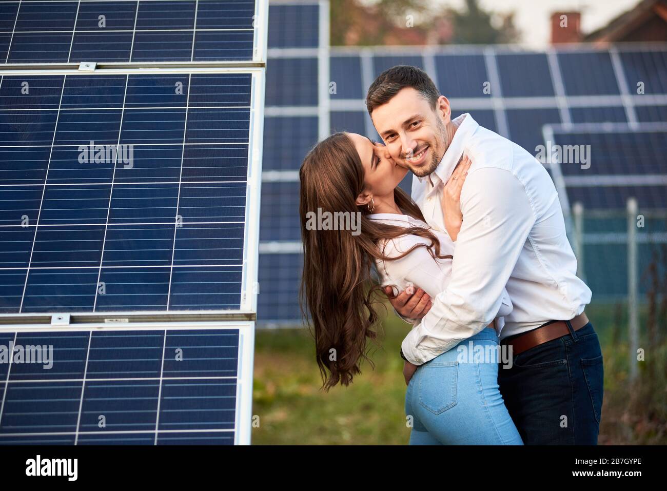 Happy pair are hugging against the background of a row of solar panels at a site near the house. Girl with long hair kisses a guy. Slender people in jeans and white shirts. Solar energy concept image Stock Photo