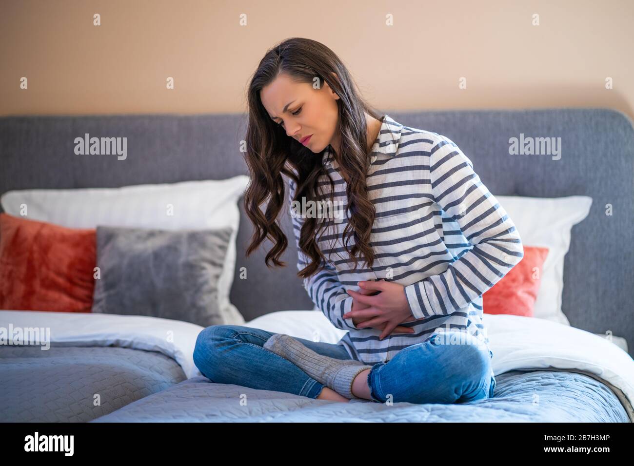 Woman is sitting on bed in her home, she has pain in stomach. Stock Photo