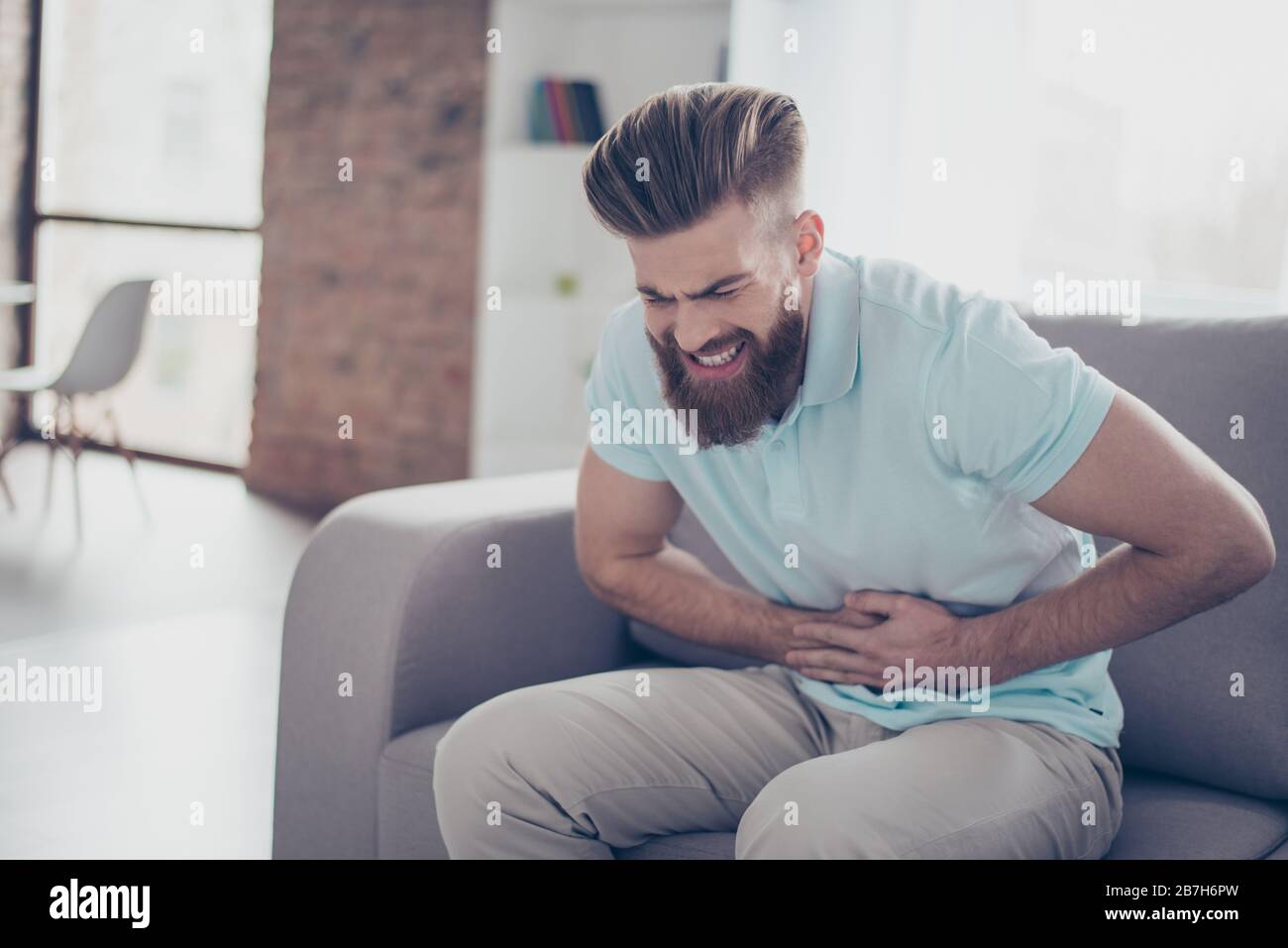 Sad young bearded man is suffering from stomach pain, ache is very hard. He is alone at home, sitting on couch Stock Photo