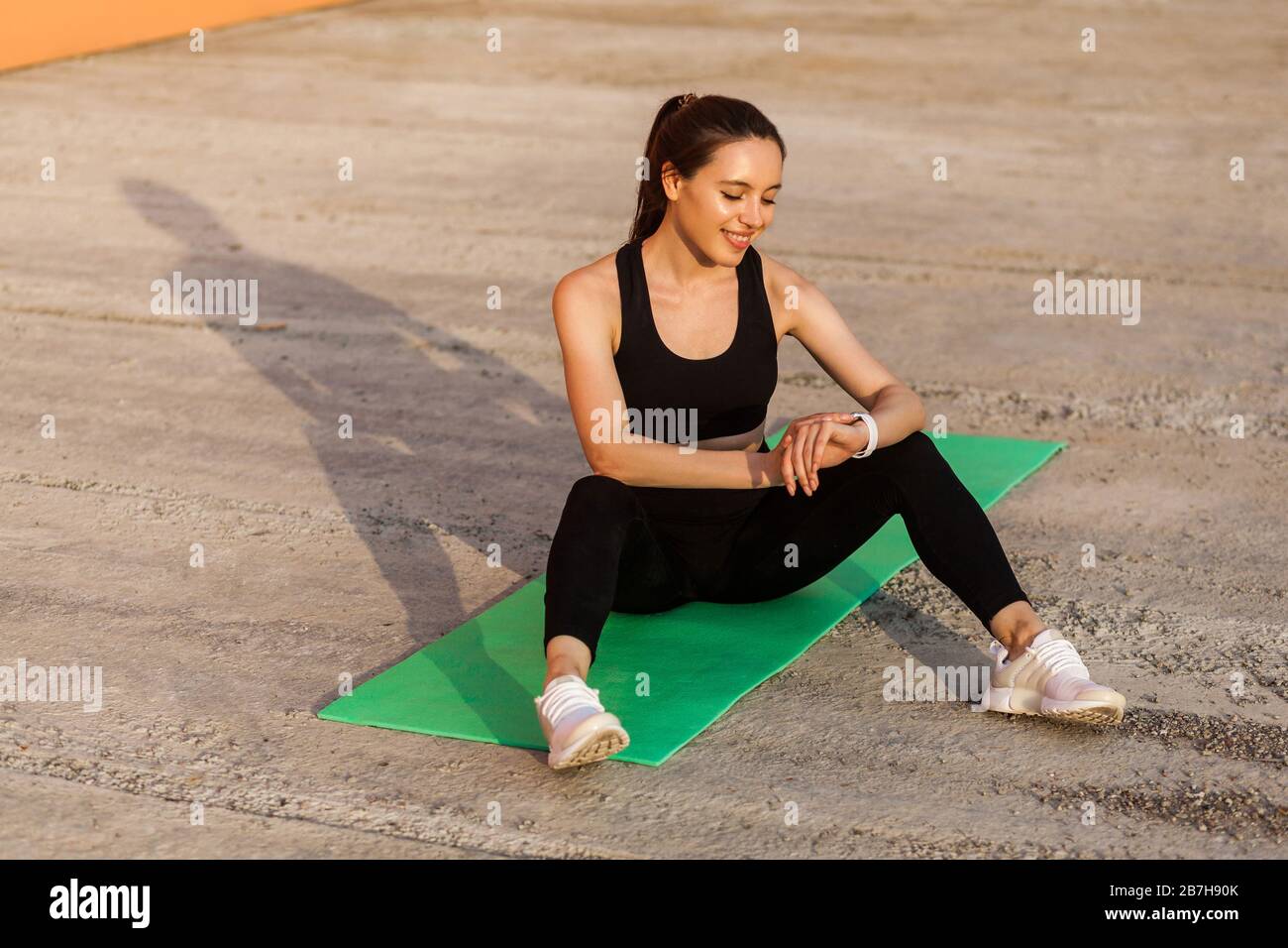 Smiling athletic pretty girl in tight sportswear, black pants and top, sitting on yoga mat after workouts, checking body health indicators on smart wa Stock Photo