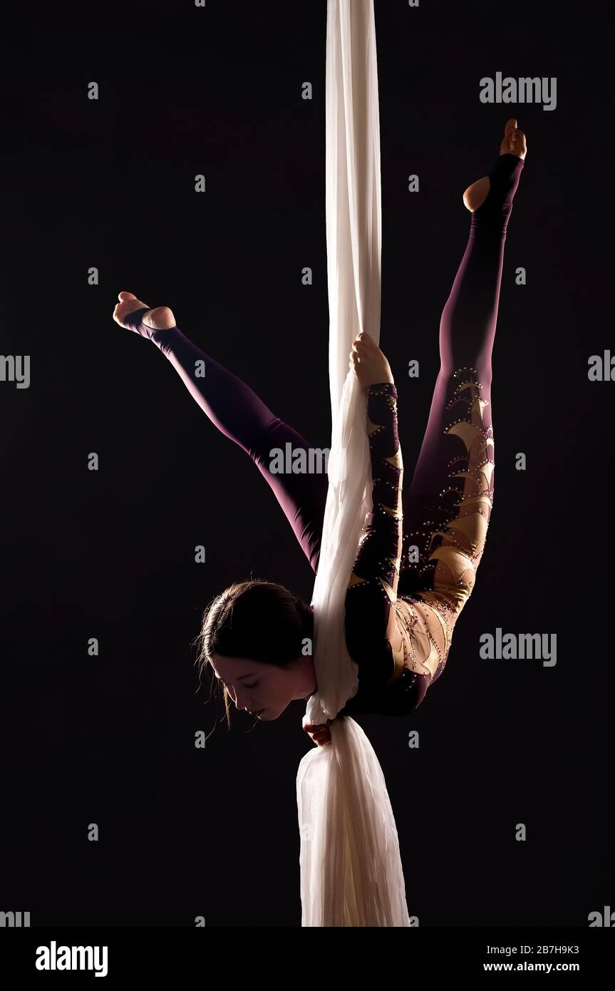 Sporty woman in a Burgundy suit  performs gymnastic and circus exercises on white silk, in the contra light. Studio shooting on a dark background. Stock Photo