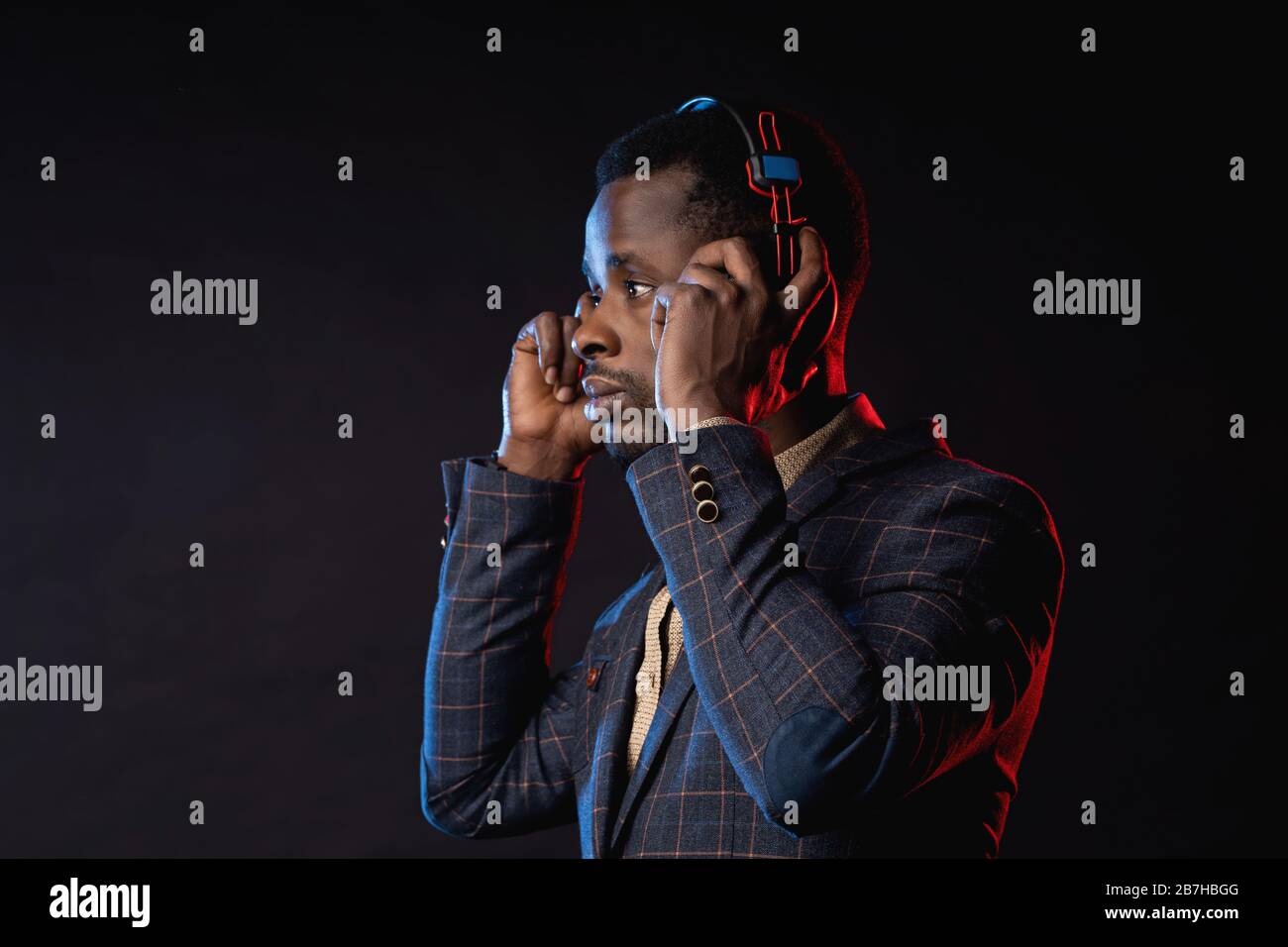 Closeup shot of african american young unshaven man listening music in headphones, looking down, posing in dark. Portrait of stylish DJ with headset, Stock Photo