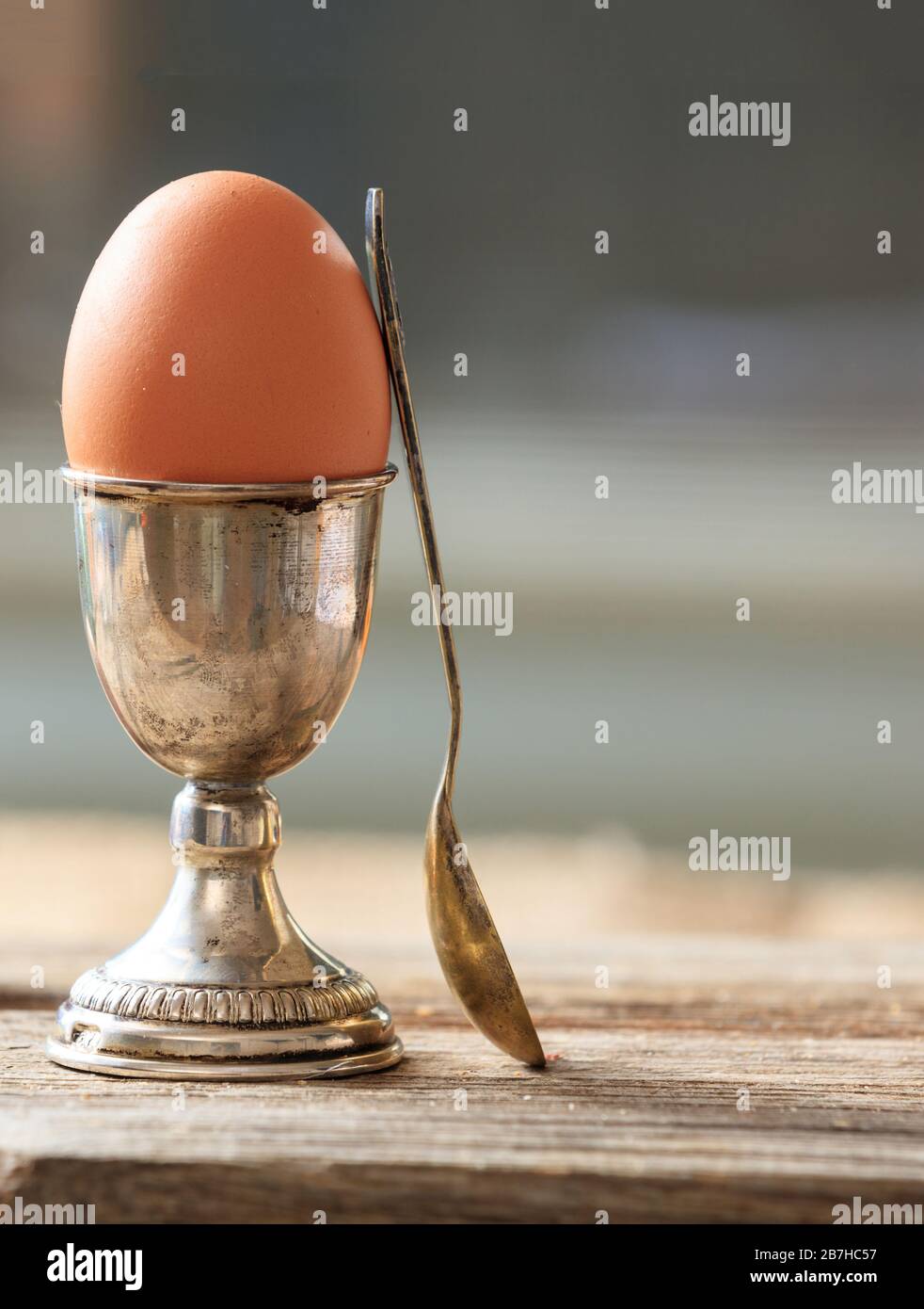 Egg in a silver eggcup and a spoon on wooden table. Vertical portrait of the set and a fresh full protein brown egg. Blur background, space. Stock Photo