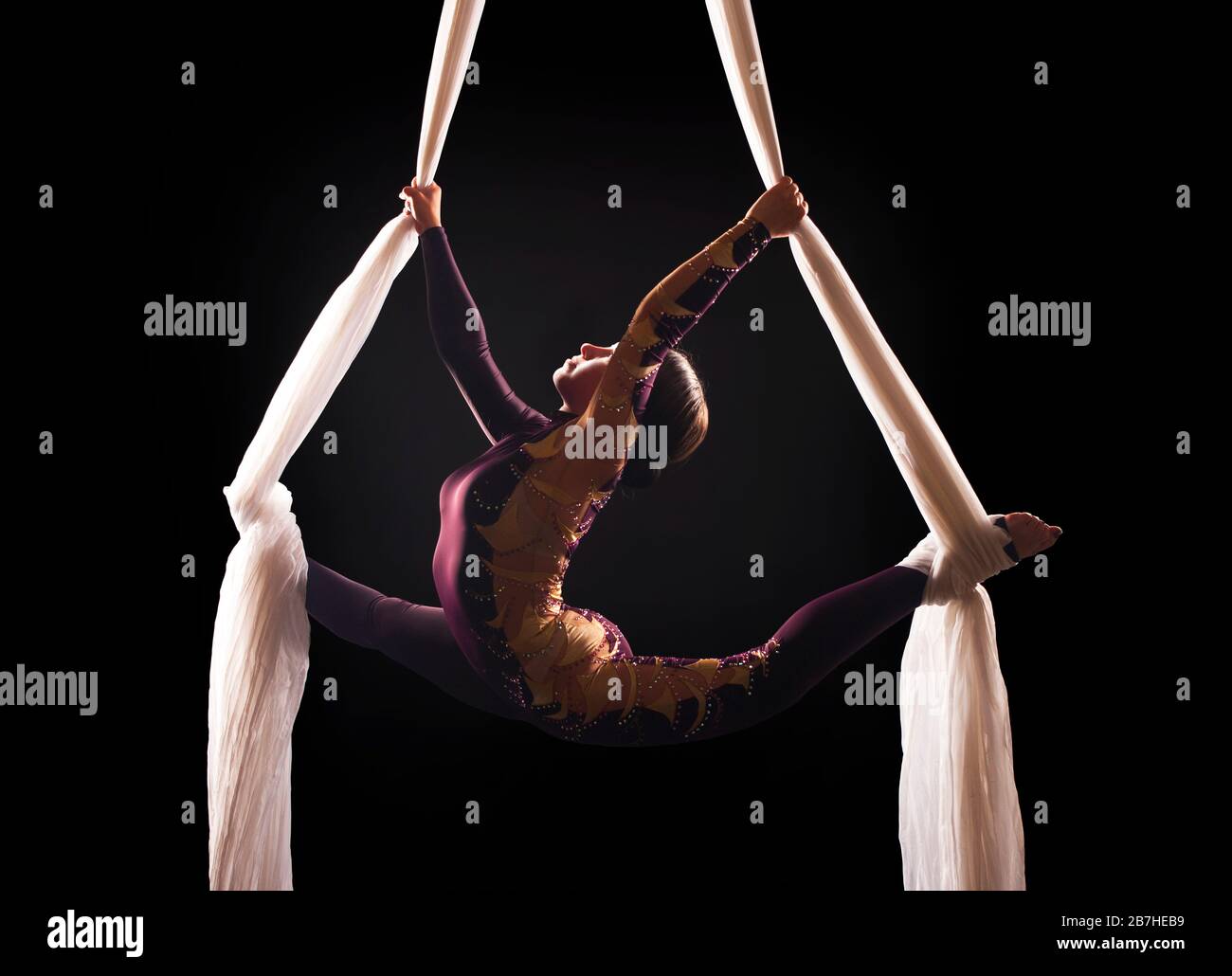 Sporty woman in a Burgundy suit  performs gymnastic and circus exercises on white silk, in the contra light. Studio shooting on a dark background. Stock Photo