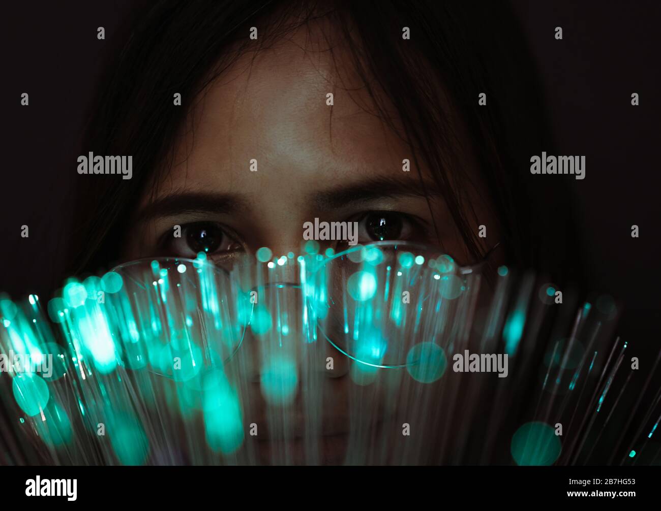 Diverse Asian girl looking through futuristic, teal coloured bokeh - Millennial woman looking at camera with advanced fibre optic technology - Next generation super fast internet and optical tech concept Stock Photo