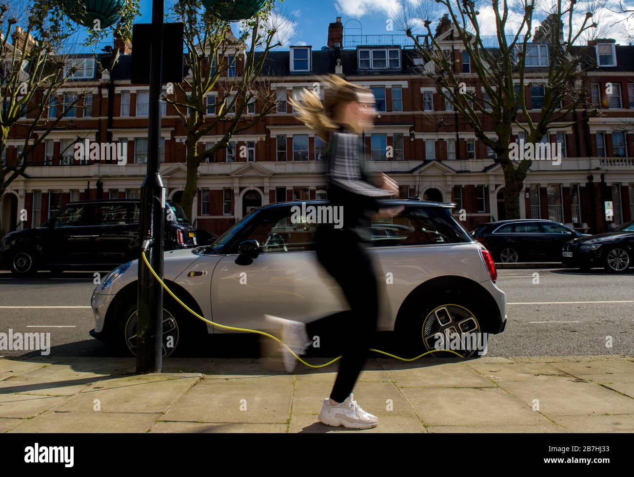 A woman runs past an electric Mini Cooper charging on Sutherland Avenue, London, which has been coined as Electric Avenue, W9, to mark that all lampposts on the street have been fully converted to SIEMENS and ubitricity Electric Vehicle charging points. PA Photo. Issue date: Tuesday March 17, 2020. The street is the first of its kind in the UK, following a SIEMENS partnership with ubitricity and Westminster Council, highlighting the thousands of newly converted lampposts across London. Photo credit should read: Anthony Upton/PA Wire Stock Photo