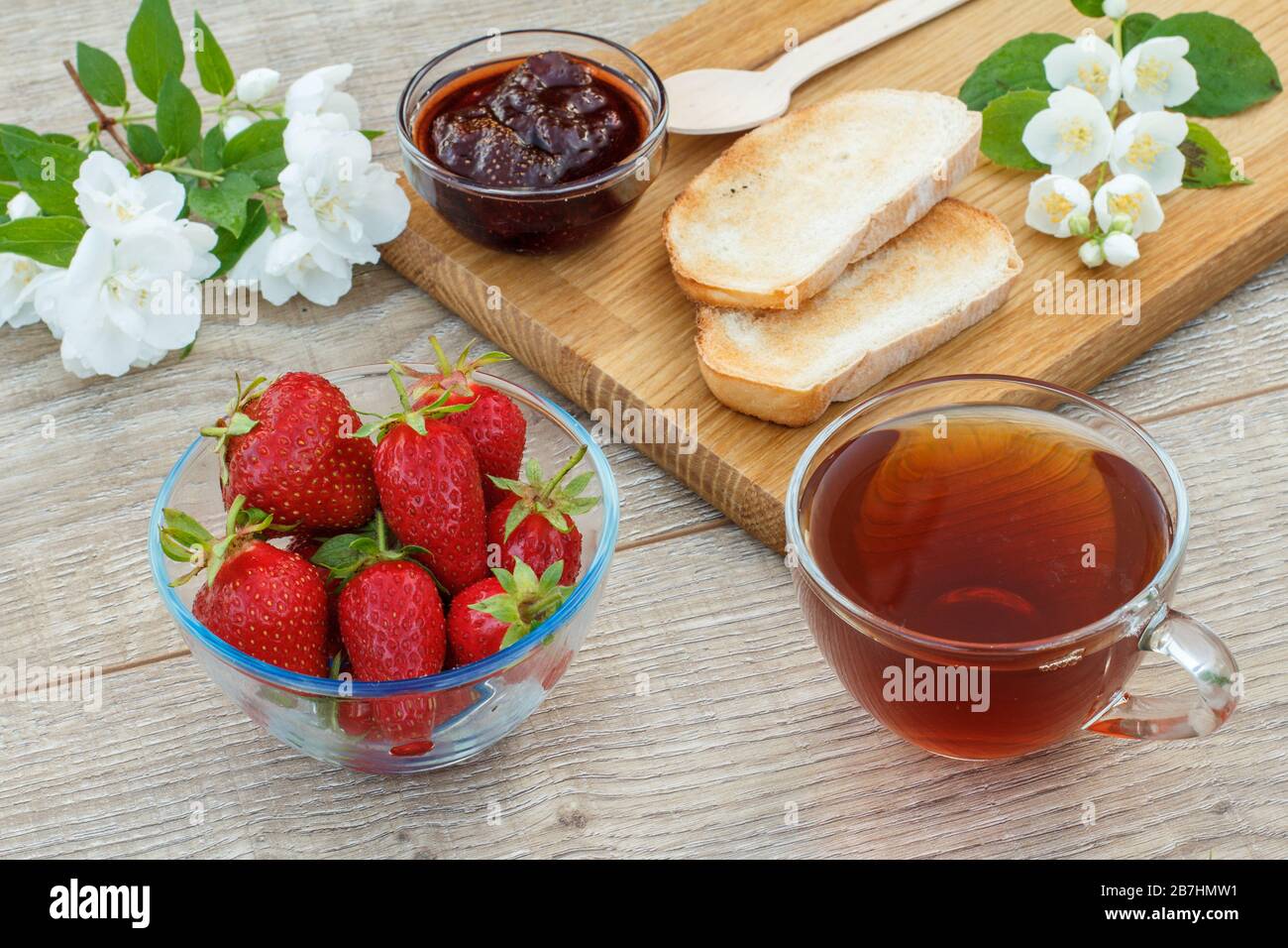 https://c8.alamy.com/comp/2B7HMW1/glass-cup-of-tea-homemade-strawberry-jam-bread-on-wooden-cutting-board-and-fresh-strawberries-spoon-and-white-jasmine-flowers-on-wooden-background-2B7HMW1.jpg