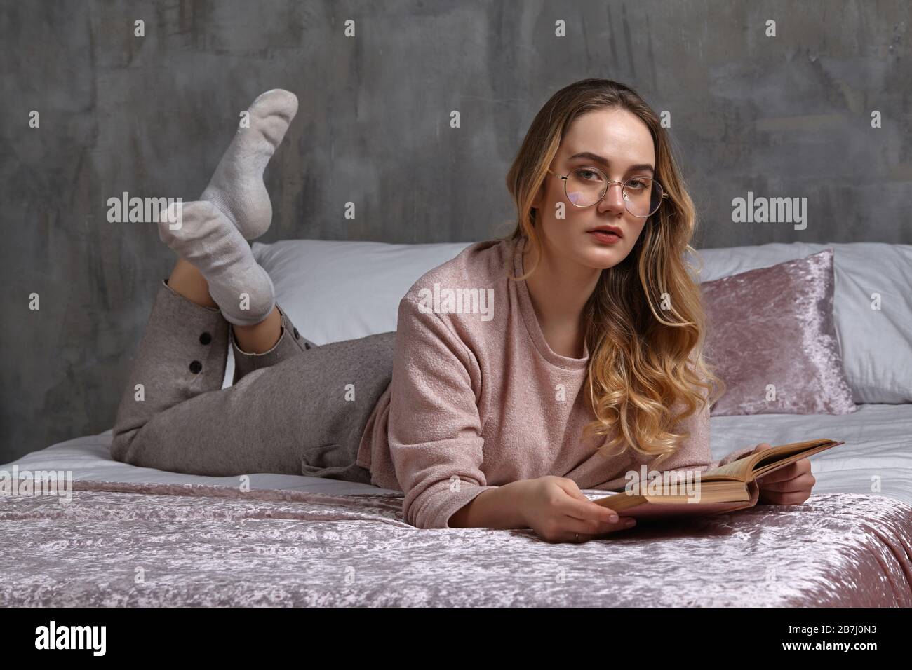 Blonde woman in glasses, casual clothing. She is looking at you, holding book, laying on bed in bedroom. Student, blogger, document studying. Close-up Stock Photo