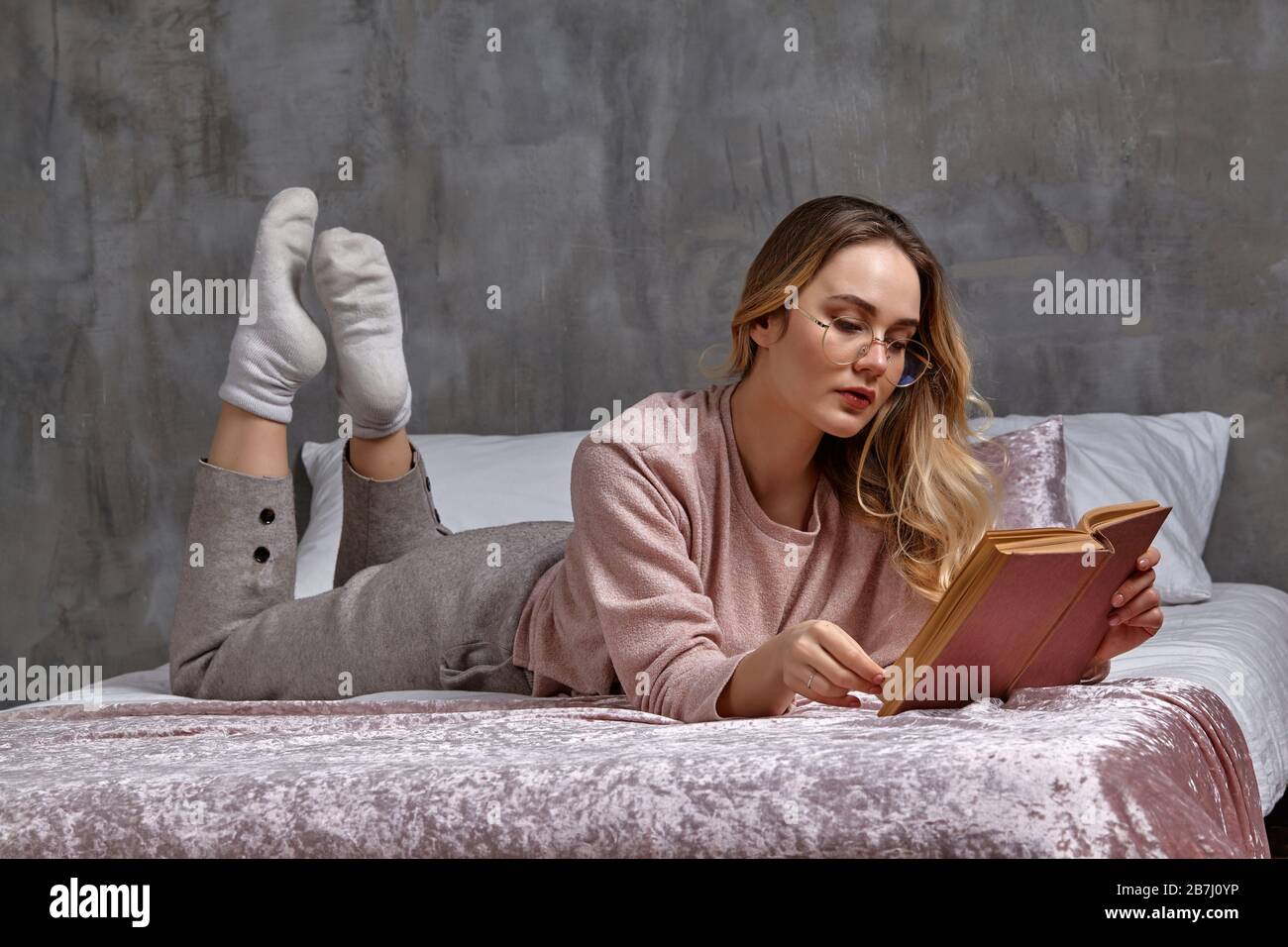Blonde woman in glasses, casual clothing. She is laying on bed and reading book in bedroom. Student, blogger. Interior with gray wall. Close-up Stock Photo