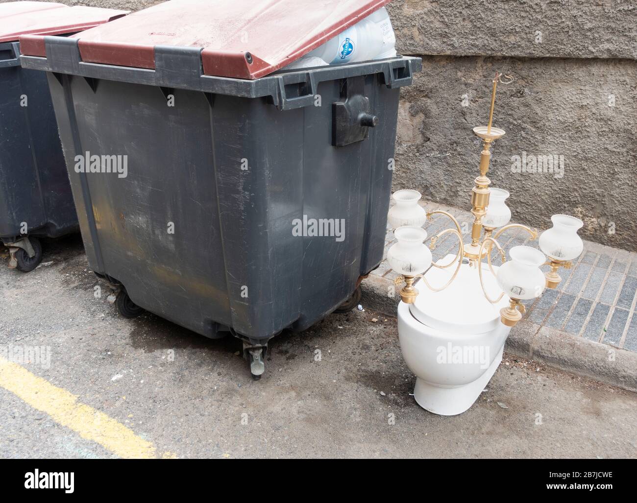 Toilet and chandelier left in street next to communal rubbish containers in street in Spain. Stock Photo