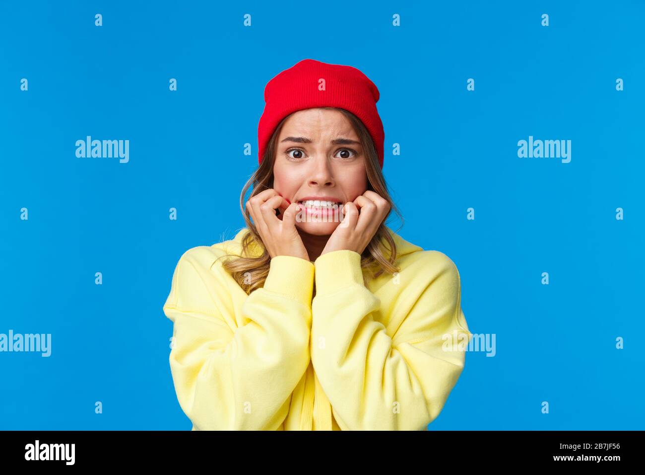 Close-up portrait of scared and insecure cute troubled caucasian girl in red beanie, biting fingernails and frowning concerned, look afraid and worrie Stock Photo