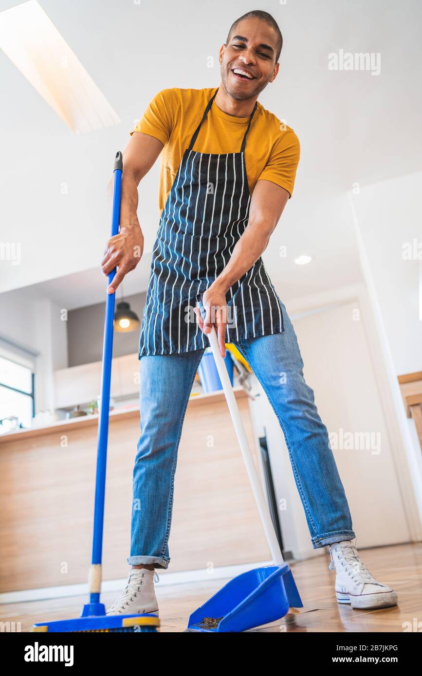 Portrait of young latin man sweeping wooden floor with broom at home. Cleaning, housework and housekeeping concept. Stock Photo