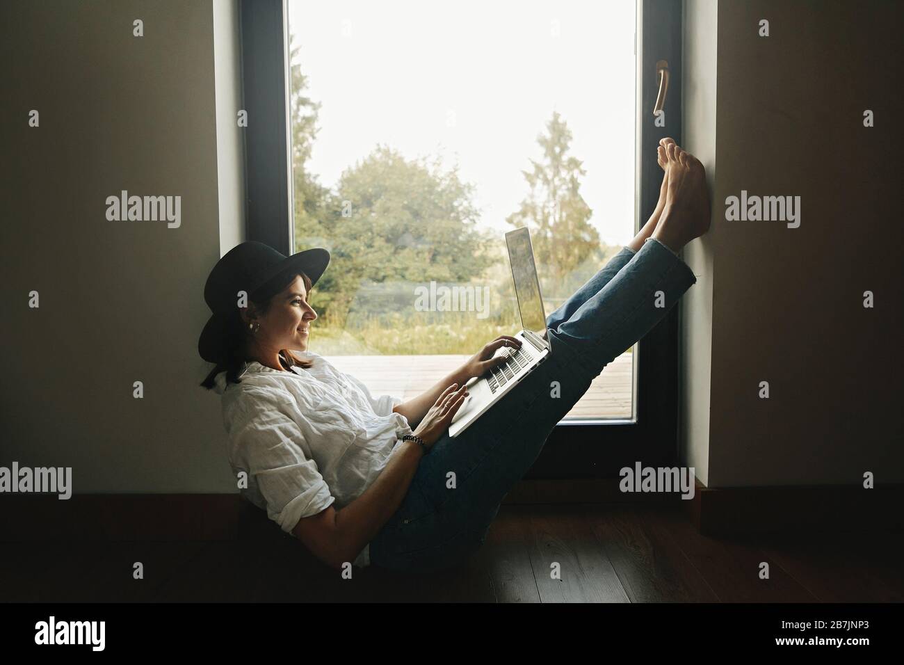 Stylish hipster girl sitting on floor with laptop at big window with view into forest. Young happy woman in hat using laptop, shopping or working onli Stock Photo