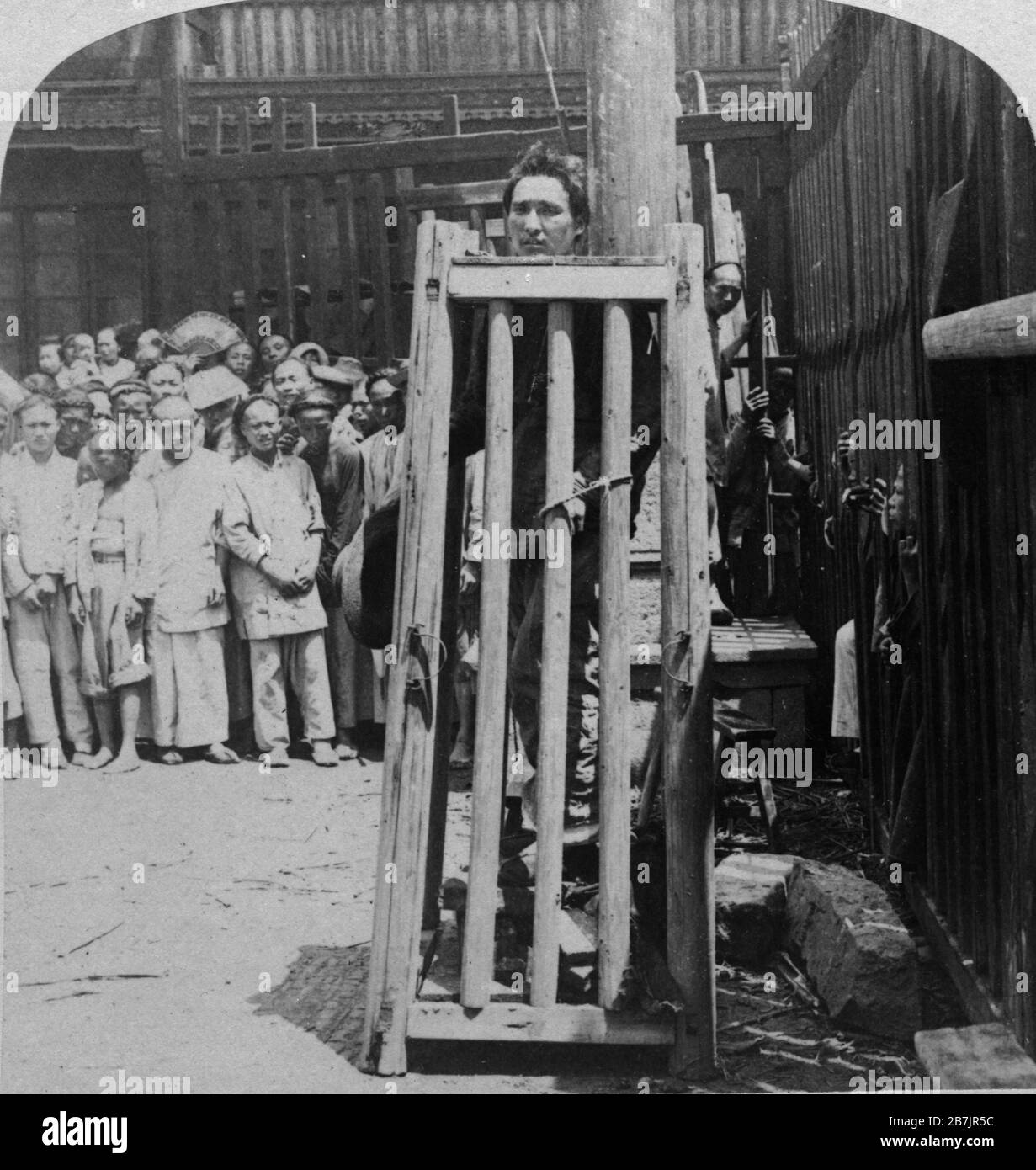 This Chinese murderer and pirate is being publicly executed in a busy thoroughfare. Cage planks fit snuggly tight around his neck. If he slips, he will strangle to death. His hands are tied to the side of his cage. He balances on a pile of flat stones and each day, one stone is removed from the pile. He will stand until he can't, or they take away the last stone. Dense crowds watched all the time. In those days, law-abiding Chinese shaved the front of their heads. Condemned criminals were held until their hair grew out.  To see my other vintage images, Search:  Prestor  vintage  China  odd Stock Photo