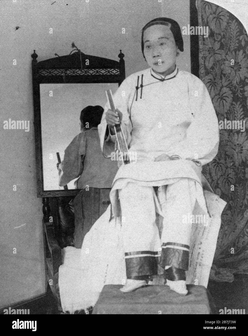 This high-caste Chinese Lady shows her dainty 'Lily feet' in 1899. She actually wears tiny silk slippers over her very tiny feet. For a thousand years, wealthy young girls' feet were cruelly broken and the four smallest toes were wrapped under the big toe. Yards of silk then bound the foot tightly. This was all to create what was considered the height of beauty for upper caste girls so they could attract the best possible husband later. The binding was socially encouraged while it was also extremely painful.   To see my related vintage images, Search:  Prestor  vintage  China  woman Stock Photo
