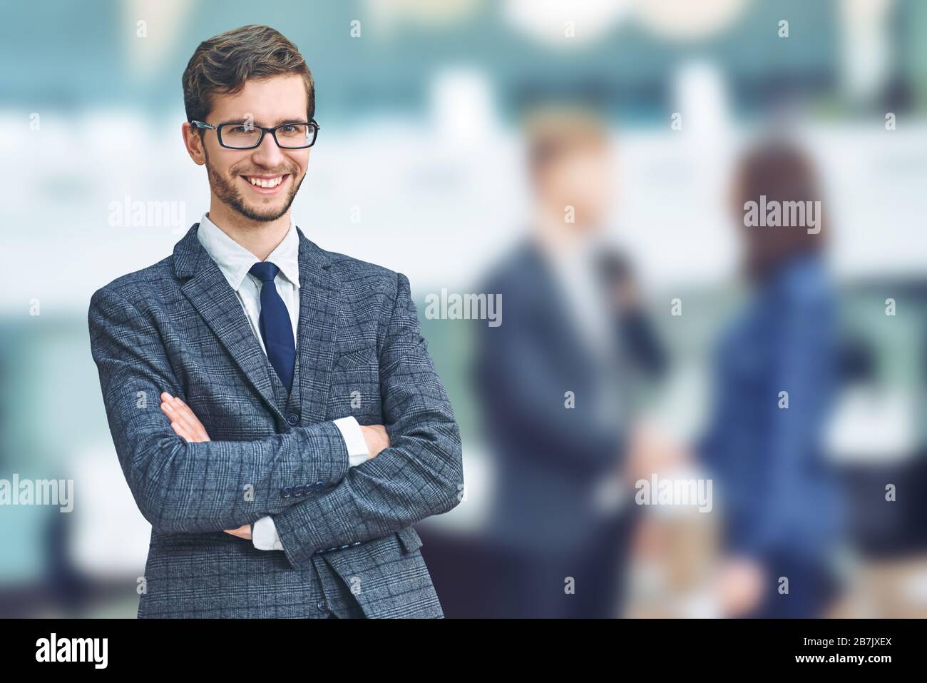 Confident and successful businessman. Cheerful young caucasian man in formalwear keeping arms crossed and smiling while his colleagues standing on Stock Photo