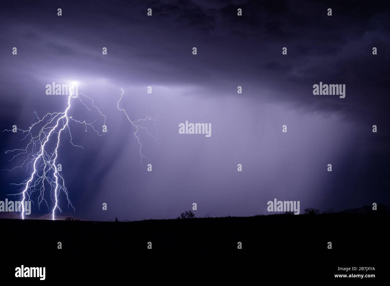 A bright cloud-to-ground lightning bolt strike illuminates a mountain as a strong thunderstorm drops heavy rain near Gila Bend, Arizona Stock Photo