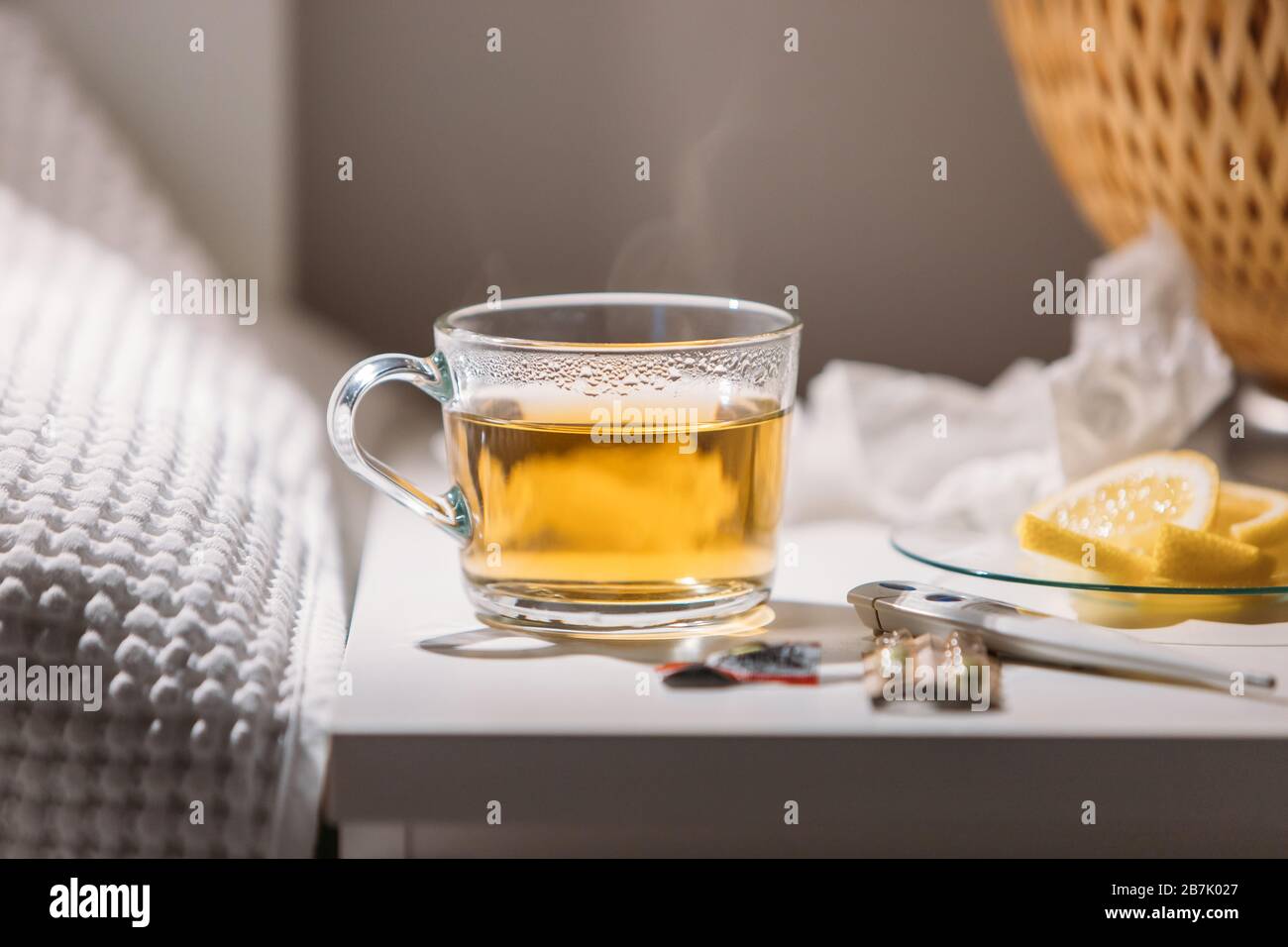 Influenza, flu season, cold concept. Close up view of cup of hot green tea, pills, digital thermometer and saucer with lemon slices on bedside table, Stock Photo