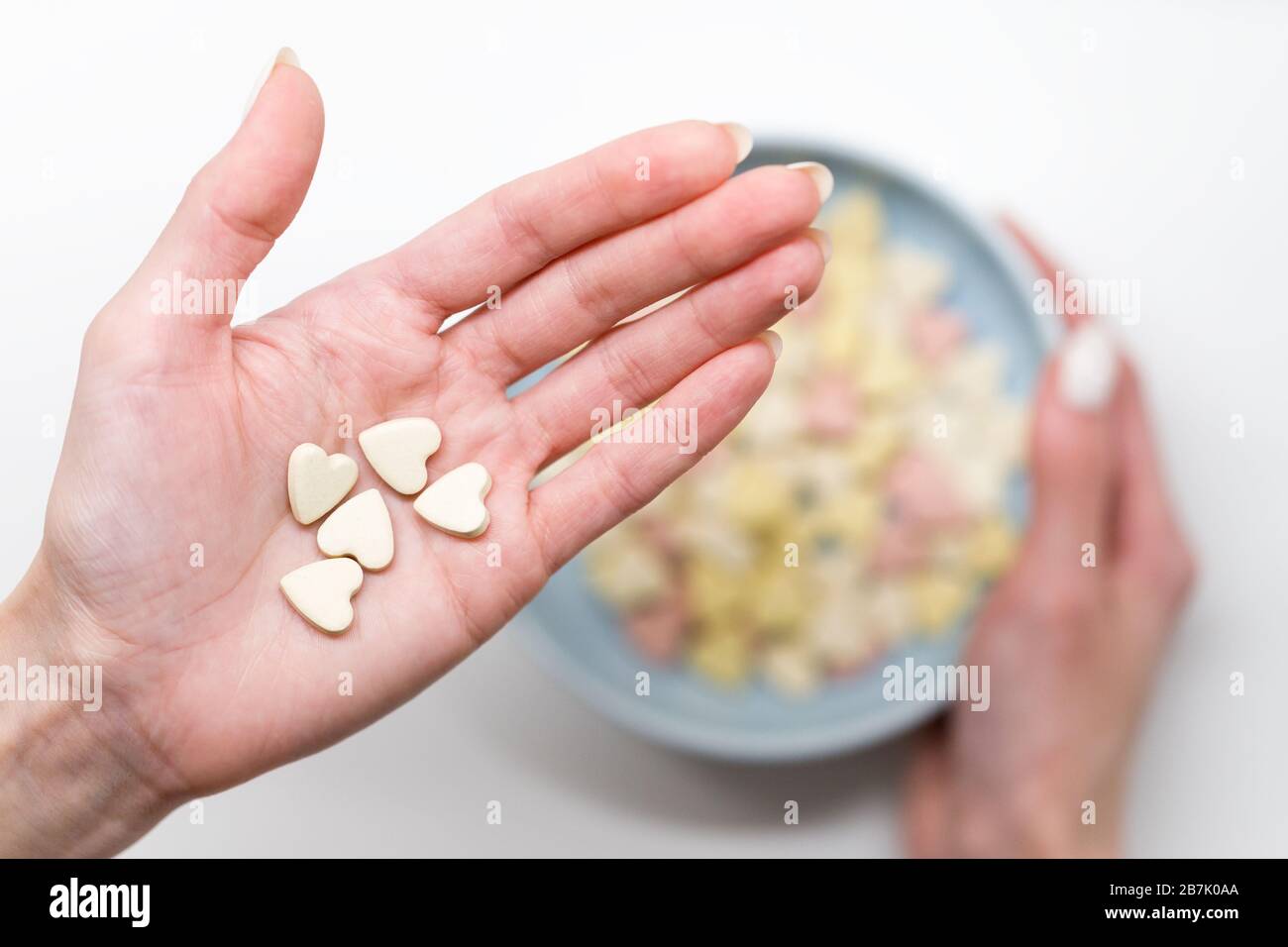 Close up of woman hold heart shaped vitamins or mineral pills on palm, blurred hand holding bowl of colorful vitamins on white background, top view. F Stock Photo