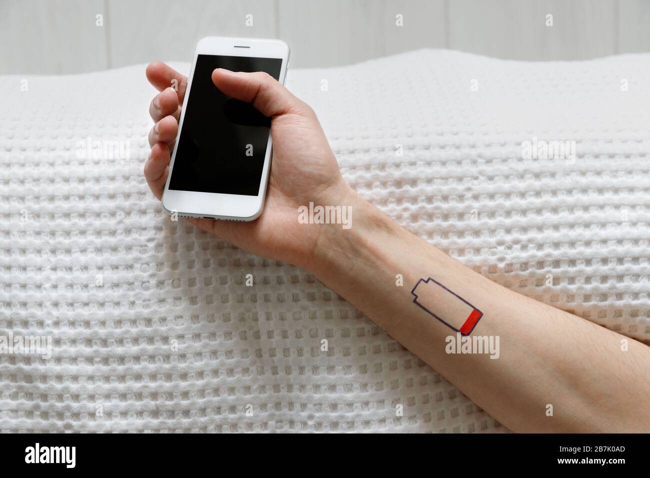 Low battery symbol drawn on human wrist. Exhaustion man lying on the bed, holding mobile phone in his hand, top view. Gadget smartphone dependence, ch Stock Photo