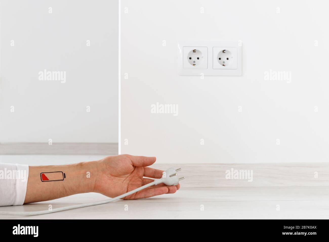 Low battery symbol drawn on human hand/wrist. Tired man lying on the floor, holding white electric plug and reaches for plug socket. Overwork, exhaust Stock Photo