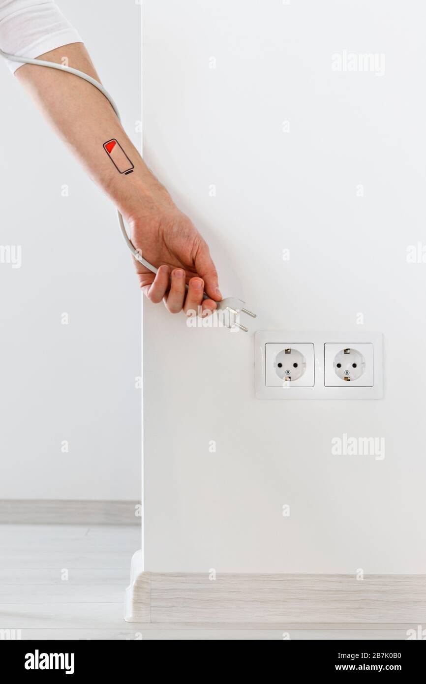 Low battery symbol drawn on human hand/wrist. Tired man holding white electric plug and reaches for plug socket. Overwork, exhausted, chronic fatigue, Stock Photo