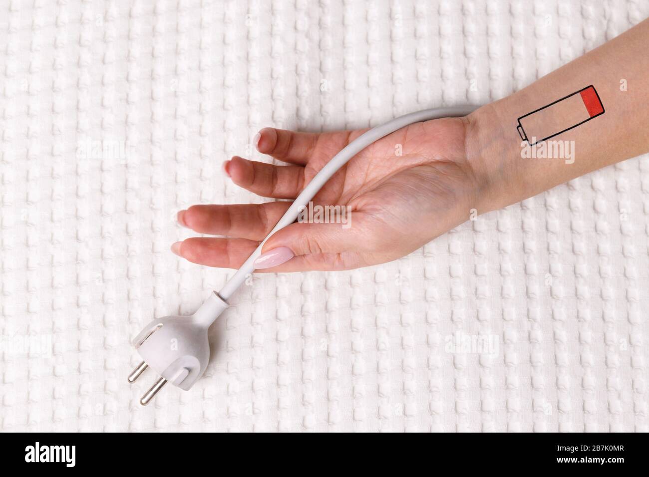 Low battery symbol drawn on human hand/wrist. Creative image of tired woman lying on bed and holding cable/white  electric plug. Overwork, exhausted, Stock Photo