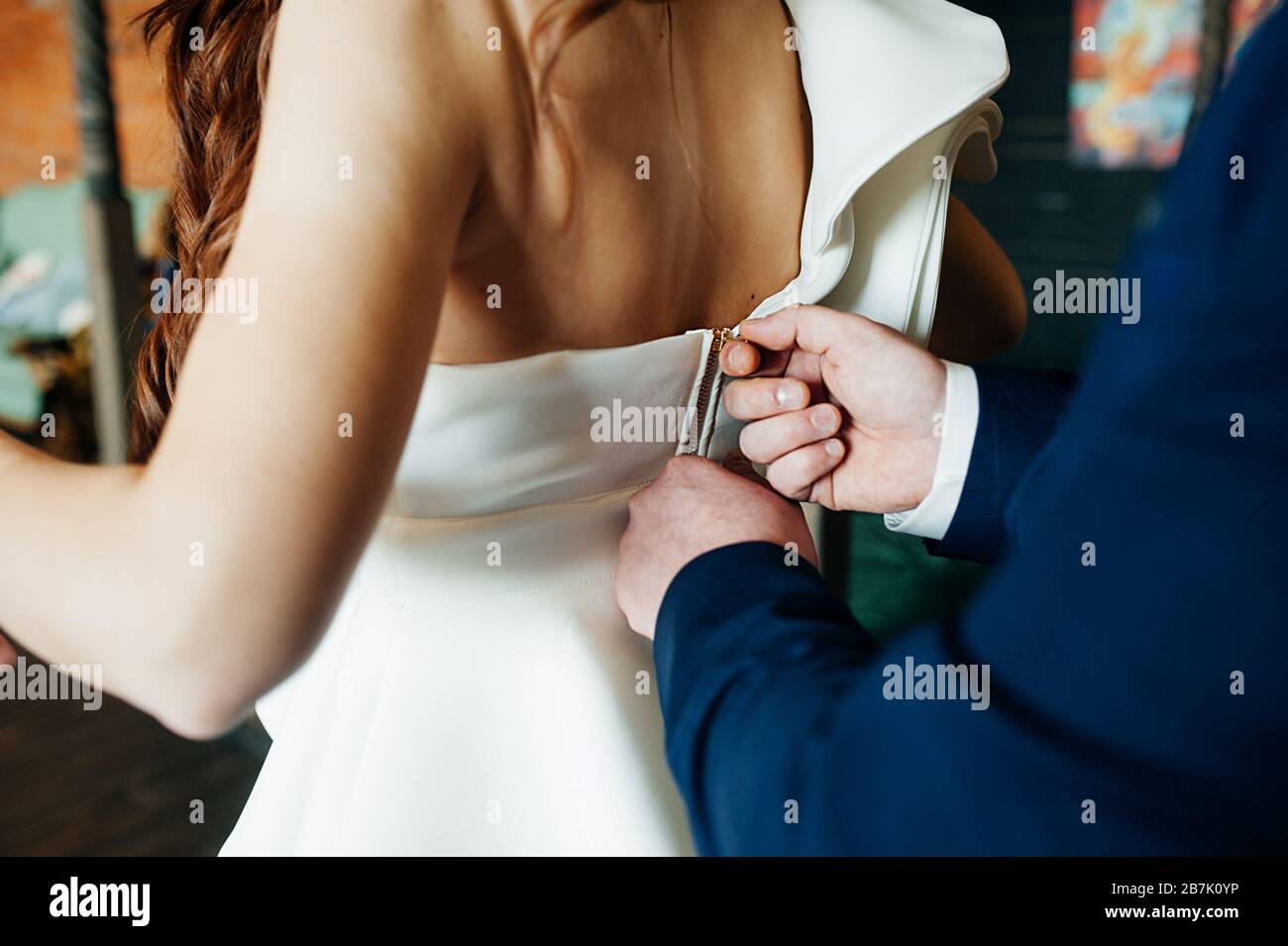 Men's rough hands in a blue suit fasten the gold zipper of a white dress on a woman's elegant back. no faces Stock Photo