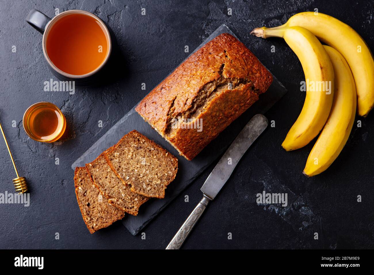 Banana, coconut bread, cake with cup of tea on slate board. Dark stone background. Copy space. Top view. Stock Photo