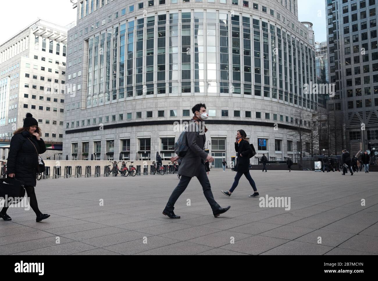 A man wearing a mask walks across Reuters Plaza in Canary Wharf, London, the day after Prime Minister Boris Johnson called on people to stay away from pubs, clubs and theatres, work from home if possible and avoid all non-essential contacts and travel in order to reduce the impact of the coronavirus pandemic. Stock Photo