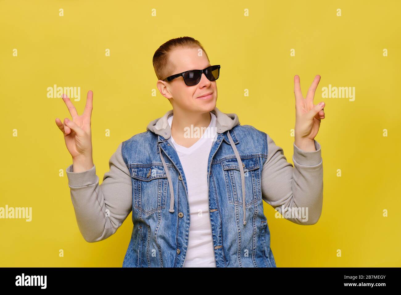 Stylish Caucasian man in a jeans and sunglasses on a yellow background. Hands abstractly shows the peace smiling. Close up. Stock Photo