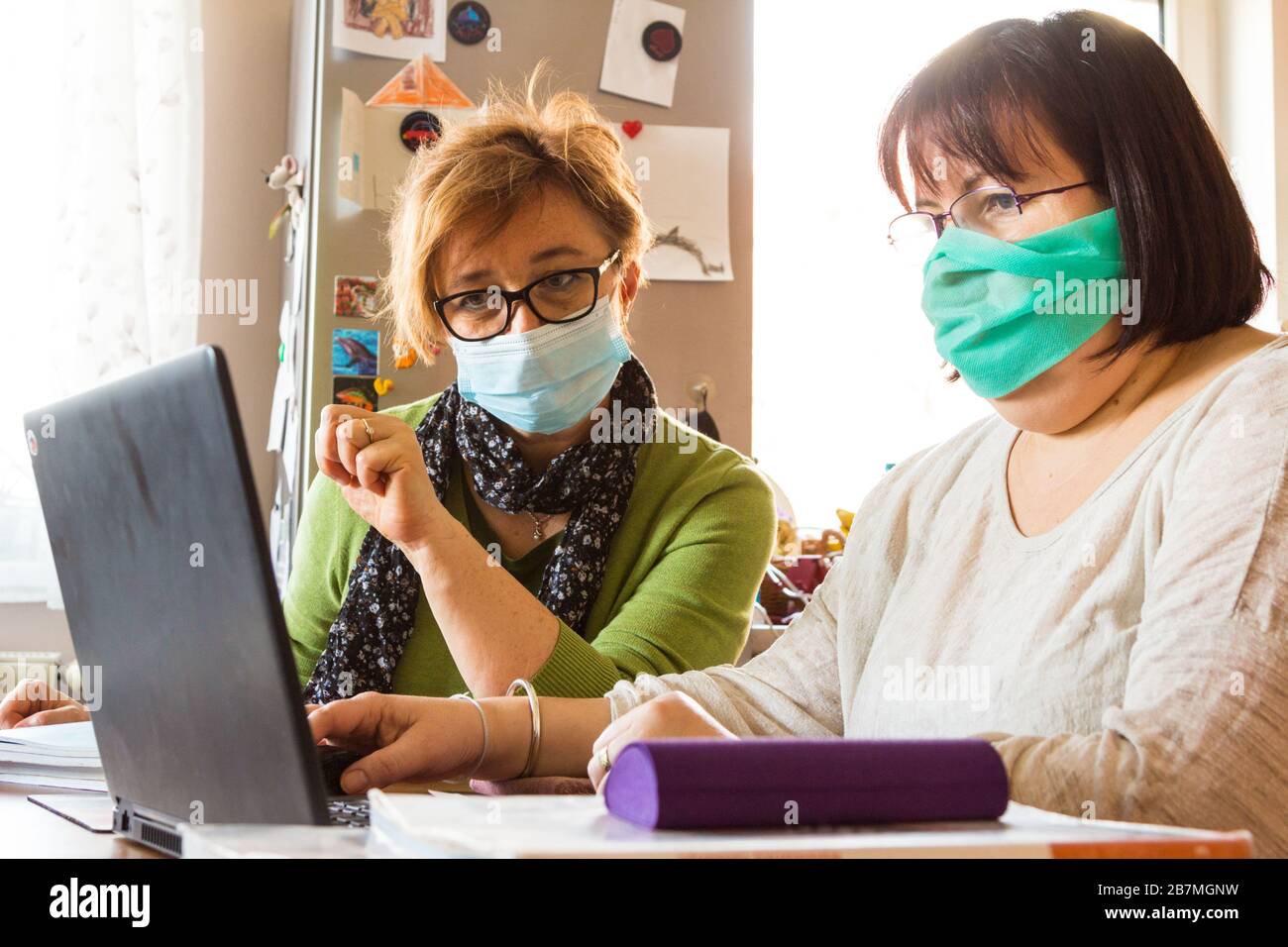 Two middle-age women teachers wearing surgical face mask discussing digital education at home during coronavirus restrictions Stock Photo