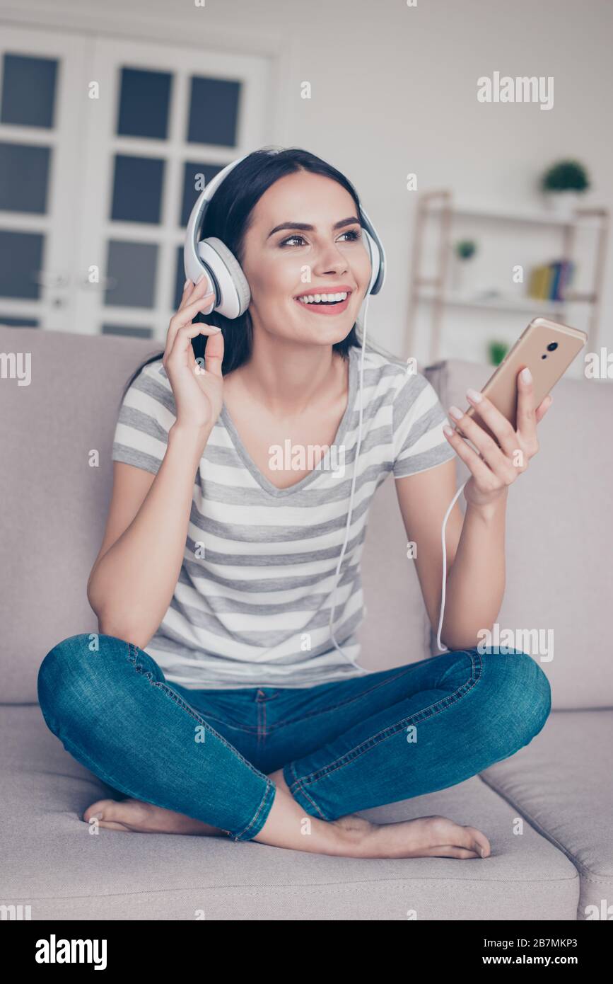 Vertical photo of young excited smiling woman sitting on a couch with crossed legs holding smartphone and listening to music Stock Photo