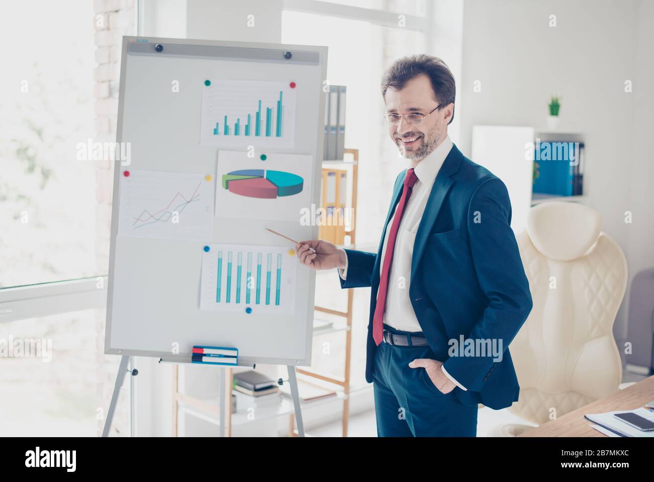 Smiling successful businessman is reporting with the flip chart in office. He is in blue suit, glasses and red tie Stock Photo