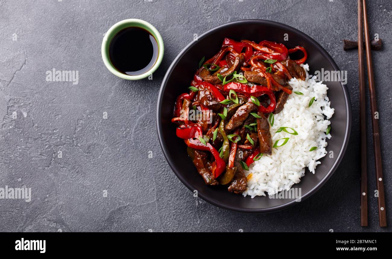 Beef and vegetables stir fry with white rice in a black bowl. Grey background. Copy space. Top view. Stock Photo