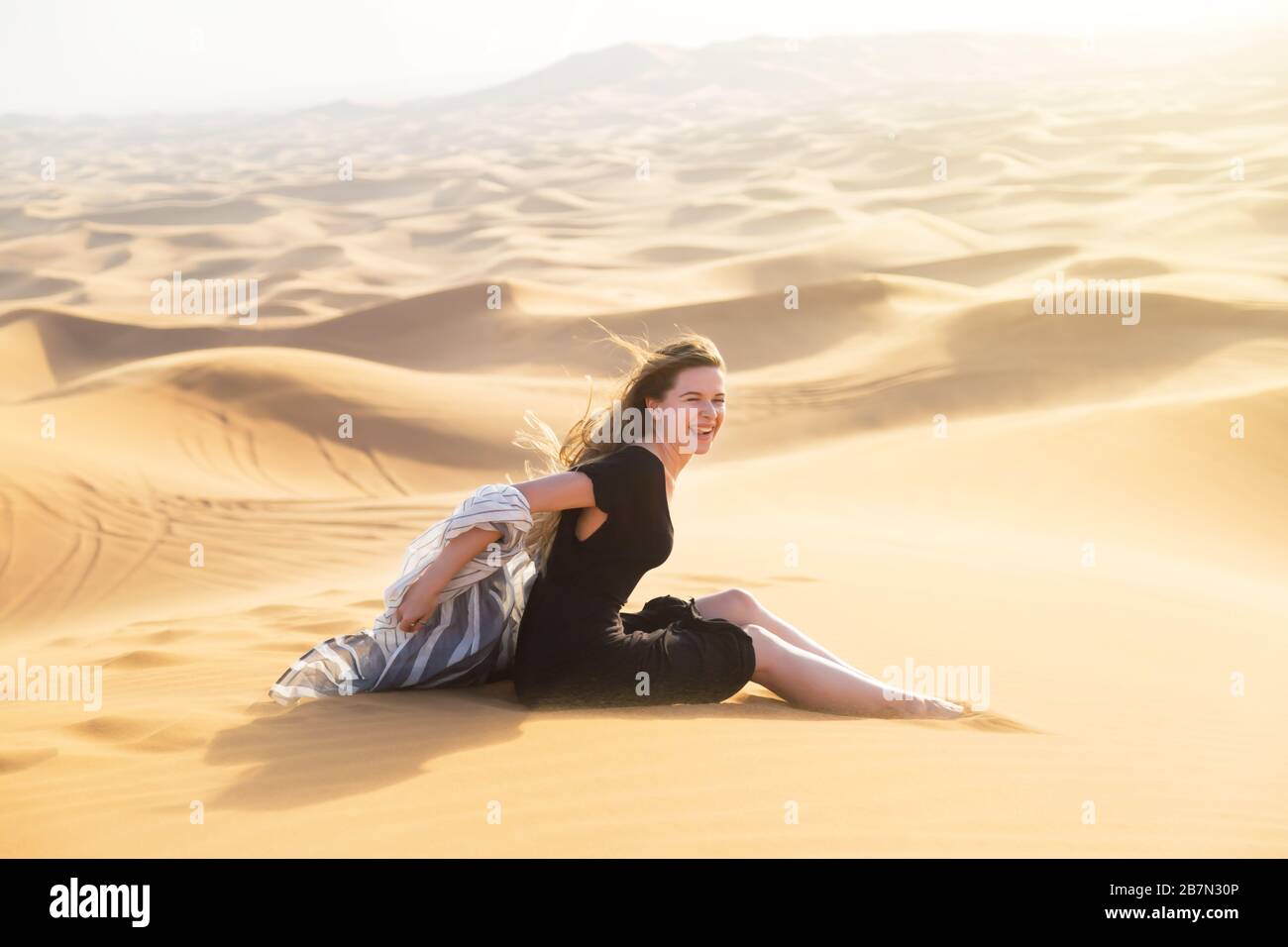 Young beautiful woman sits tiredly on the hot sand of the desert under the hot rays of the eastern sun Stock Photo