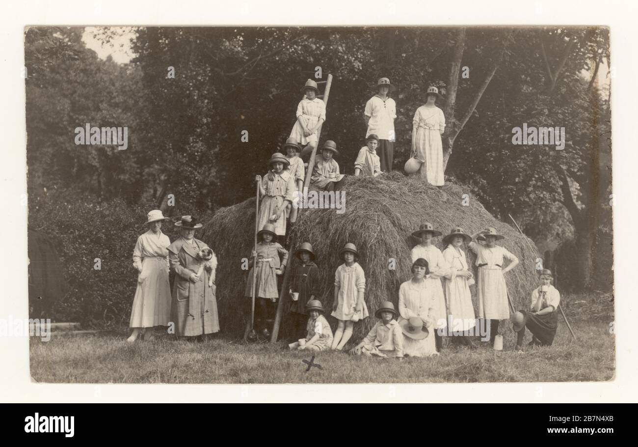 Early 1900's postcard of schoolgirls on country outing at harvest time, posing with a farmer and rakes, next to a haystack, circa 1923, U.K. Stock Photo