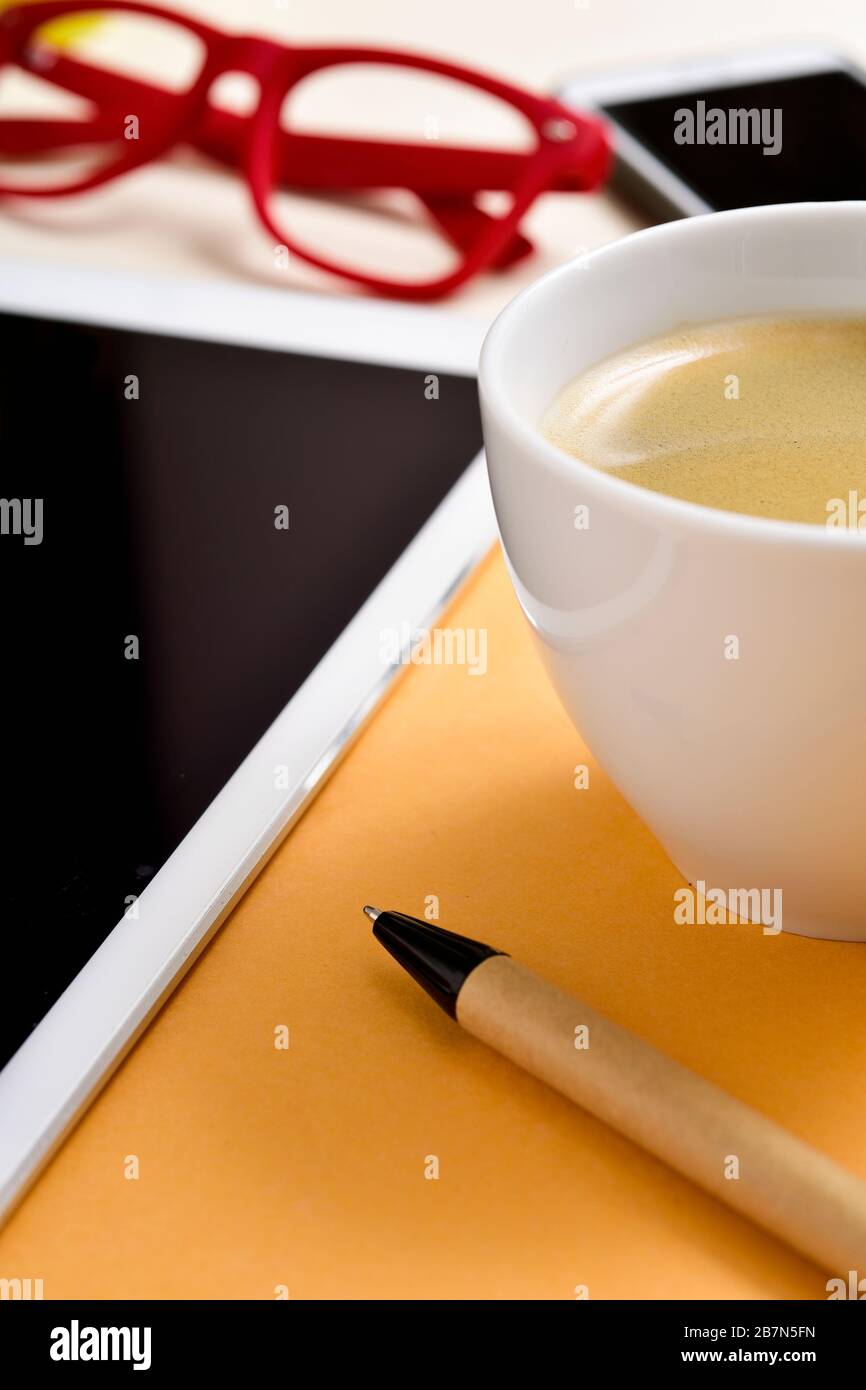 closeup of an office or home office desk full of things, such as a cup with white coffee, a pen, a tablet, a smartphone and a pair of red eyeglasses Stock Photo