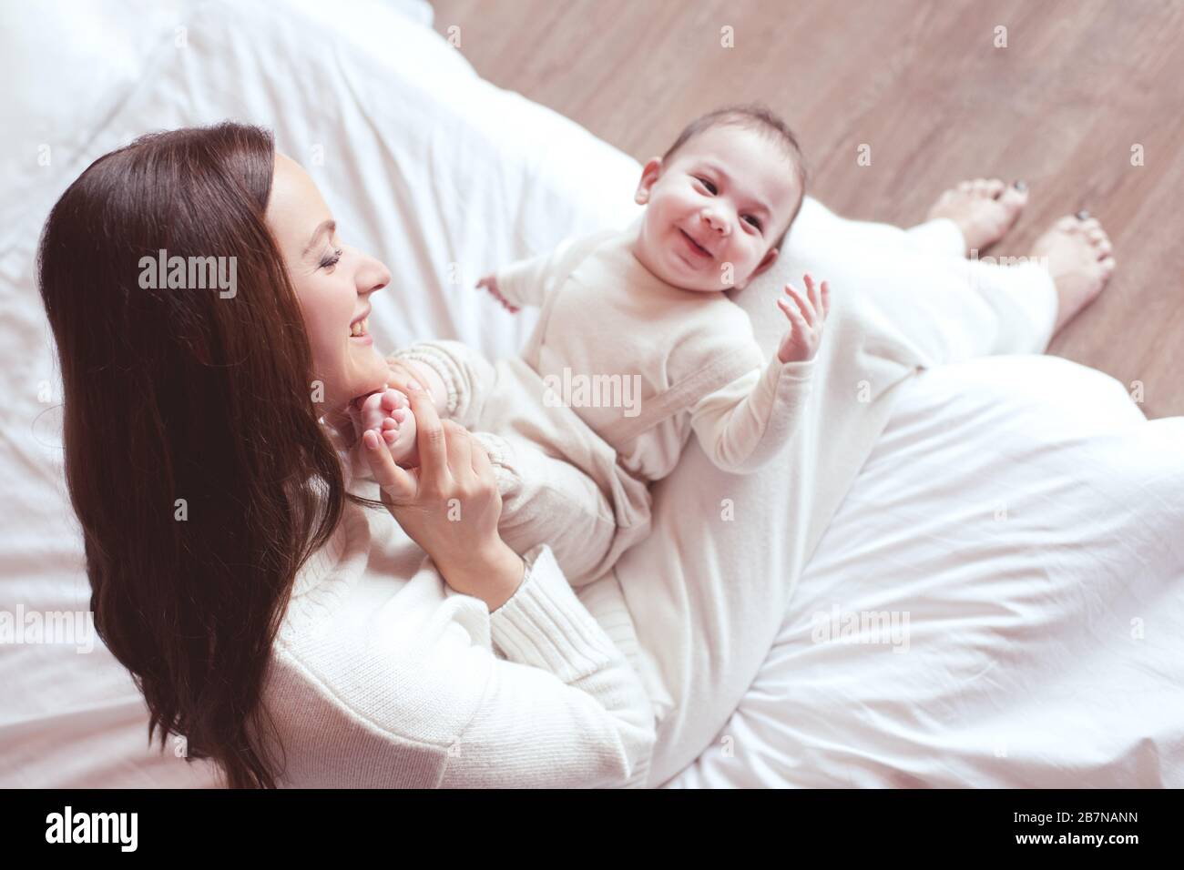 Happy woman holding adorable baby boy feet lying on her legs in room closeup. Single mother looking at her child under 1 year old. Motherhood. Good mo Stock Photo