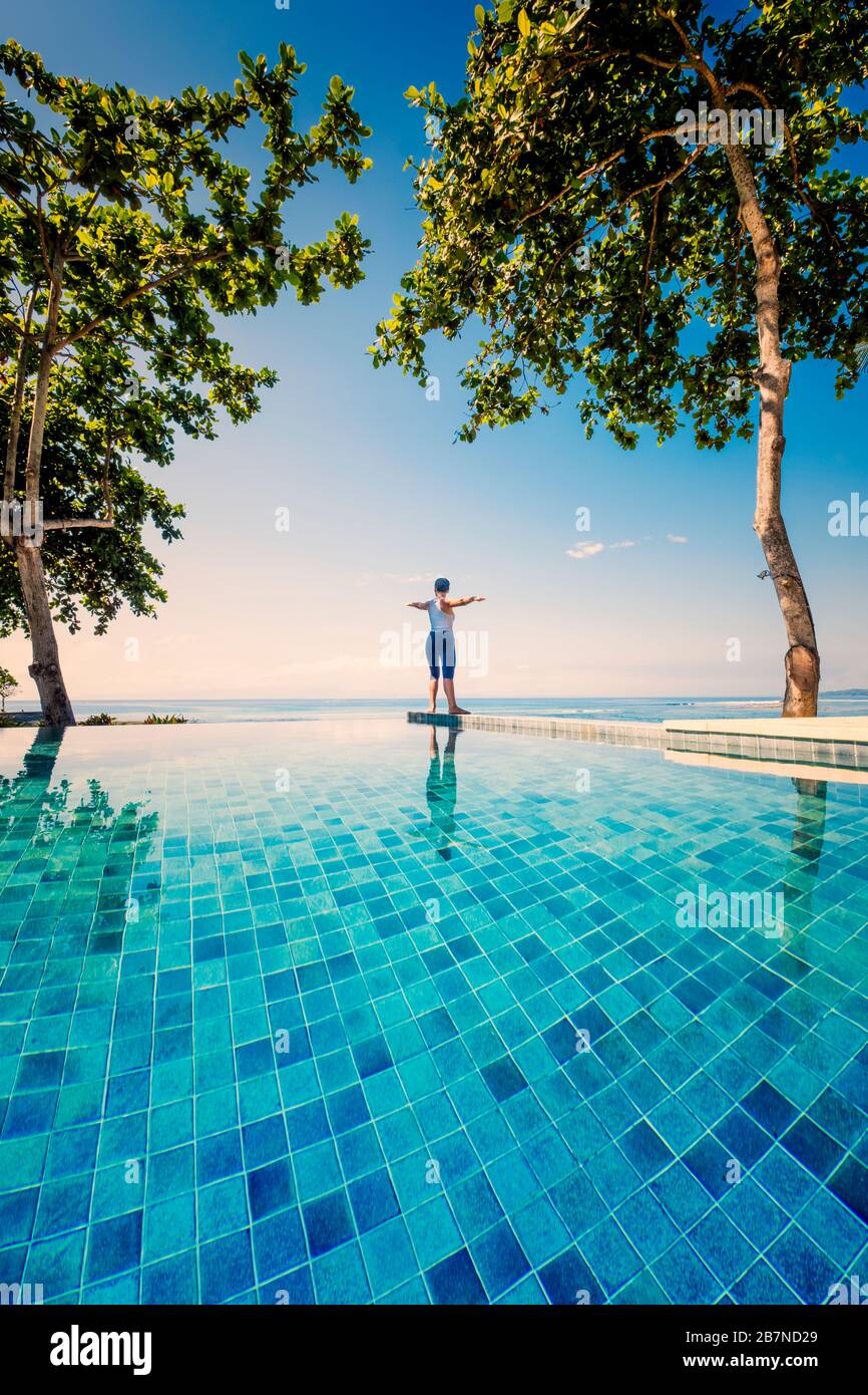 A young woman practicing yoga under palm trees next to an infinity pool Stock Photo