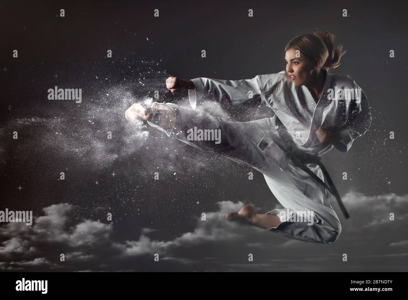 Young impudent emotional karate girl in a suit jumps up and makes a powerful blow on the background of clouds and sky. Unrestrained energy concept. Ma Stock Photo
