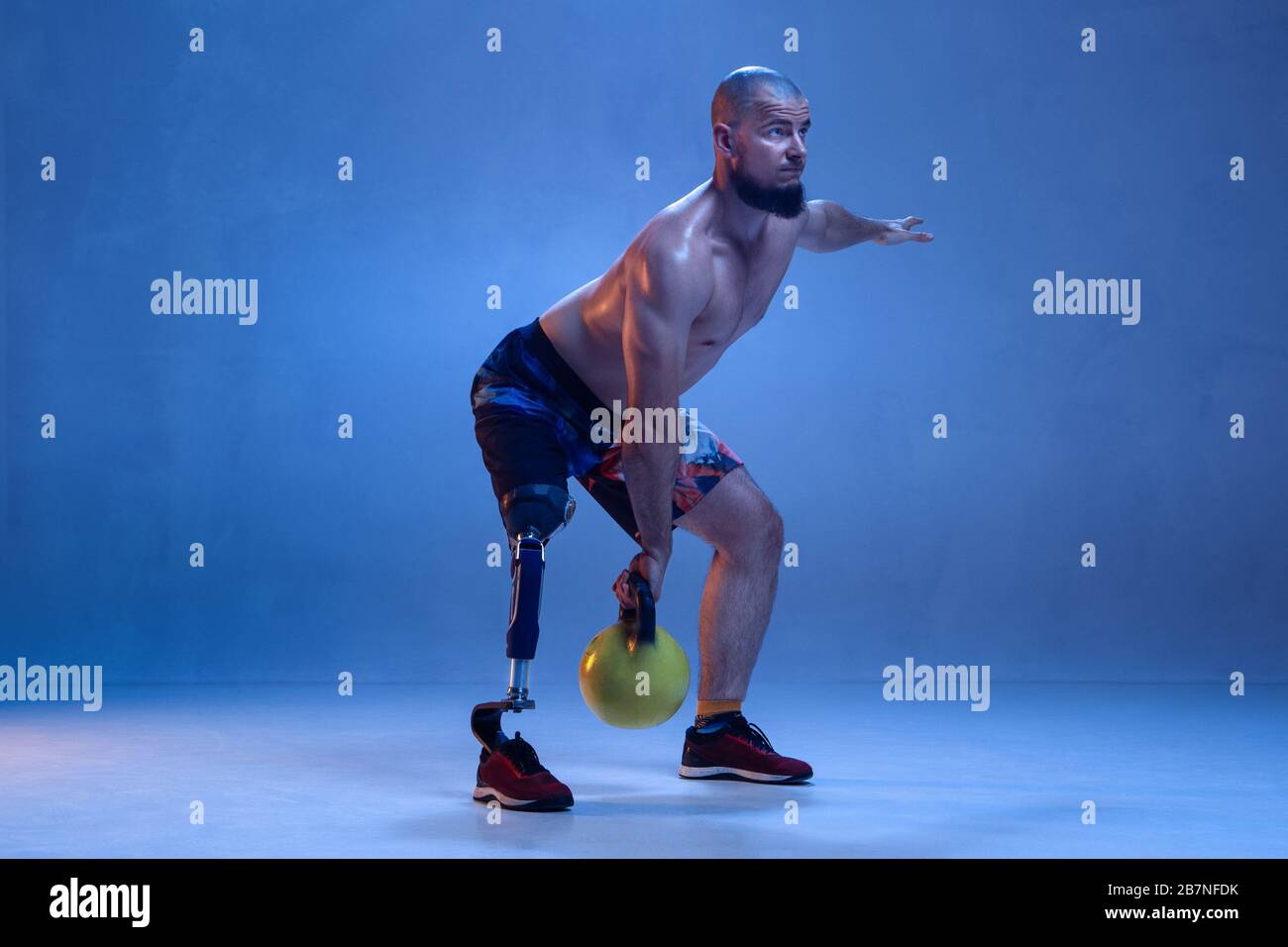Athlete with disabilities or amputee isolated on blue studio background. Professional male sportsman with leg prosthesis training with weights in neon. Disabled sport and overcoming, wellness concept. Stock Photo