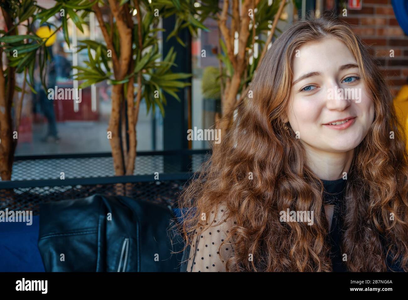 A curly-haired girl with long red hair poses in a cafe smiling at the camera. A chatty carefree student is engaged in a cafe. Stock Photo