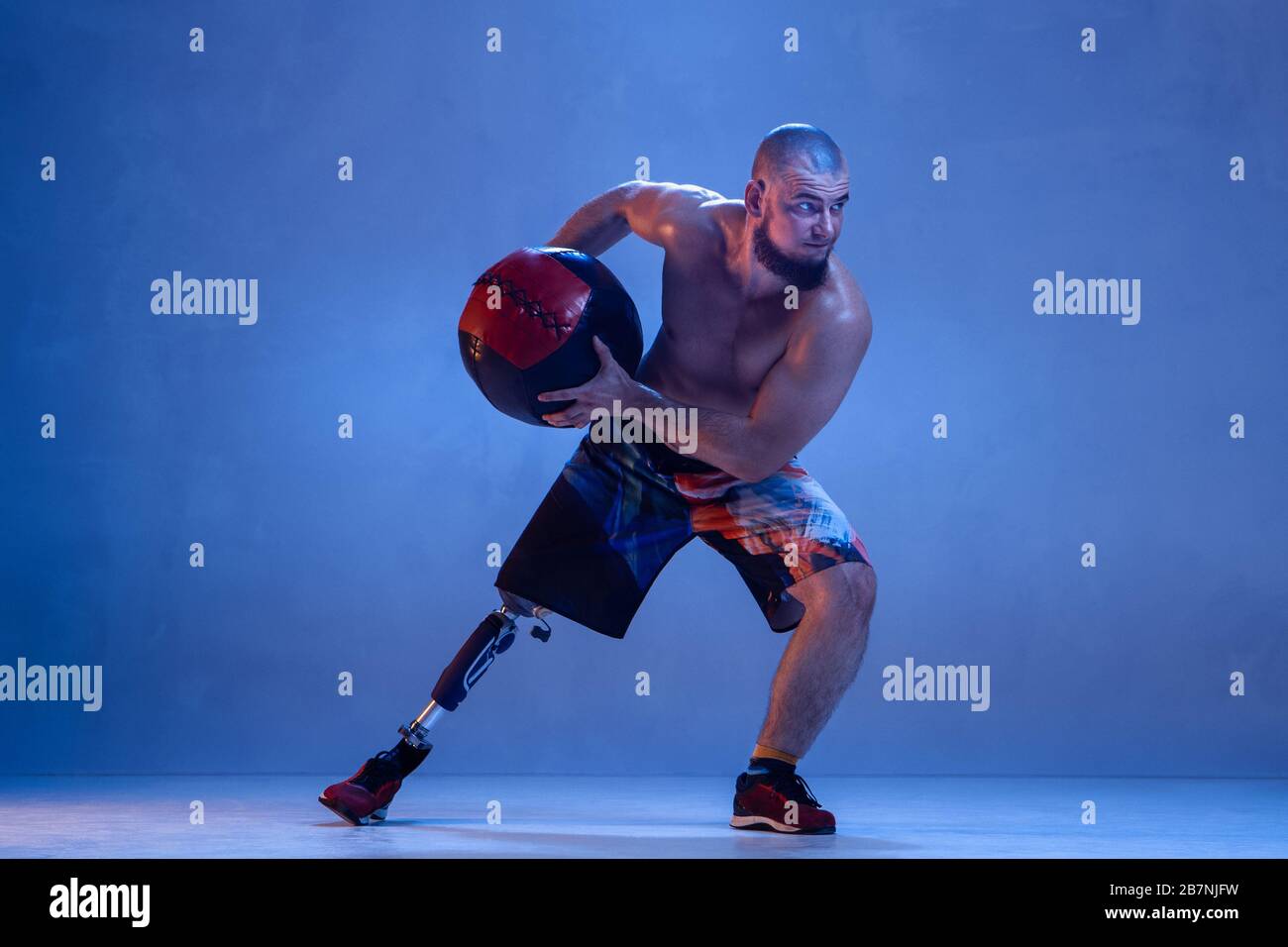 Athlete with disabilities or amputee isolated on blue studio background. Professional male sportsman with leg prosthesis training with ball in neon. Disabled sport and overcoming, wellness concept. Stock Photo