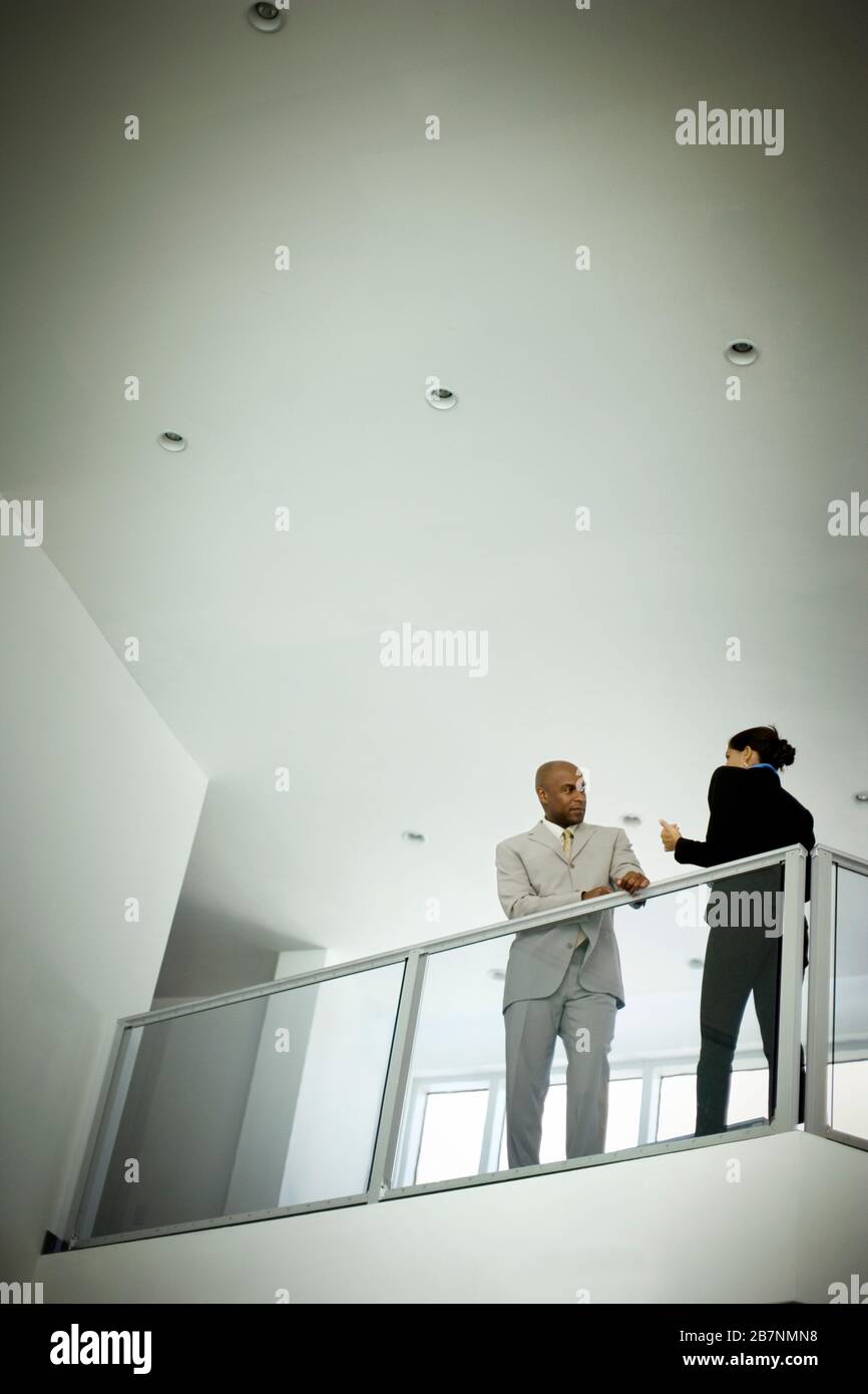 A mid-adult businessman leaning on a banister inside an office building speaking with a female colleague. Stock Photo