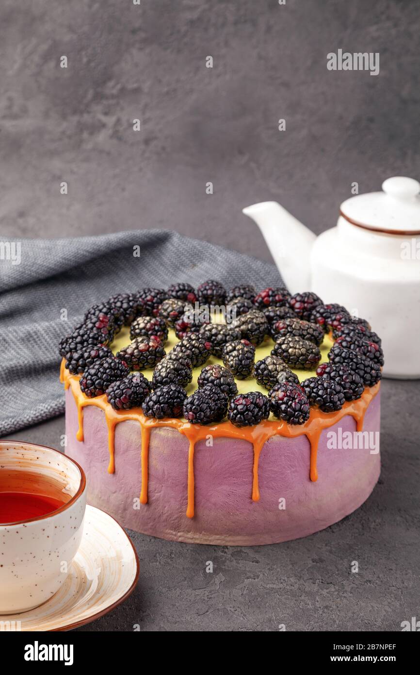 Whole cake covered with purple cream, fresh blackberries, caramel, flows down on the sides, teapot and tea cup on gray surface, vertical Stock Photo